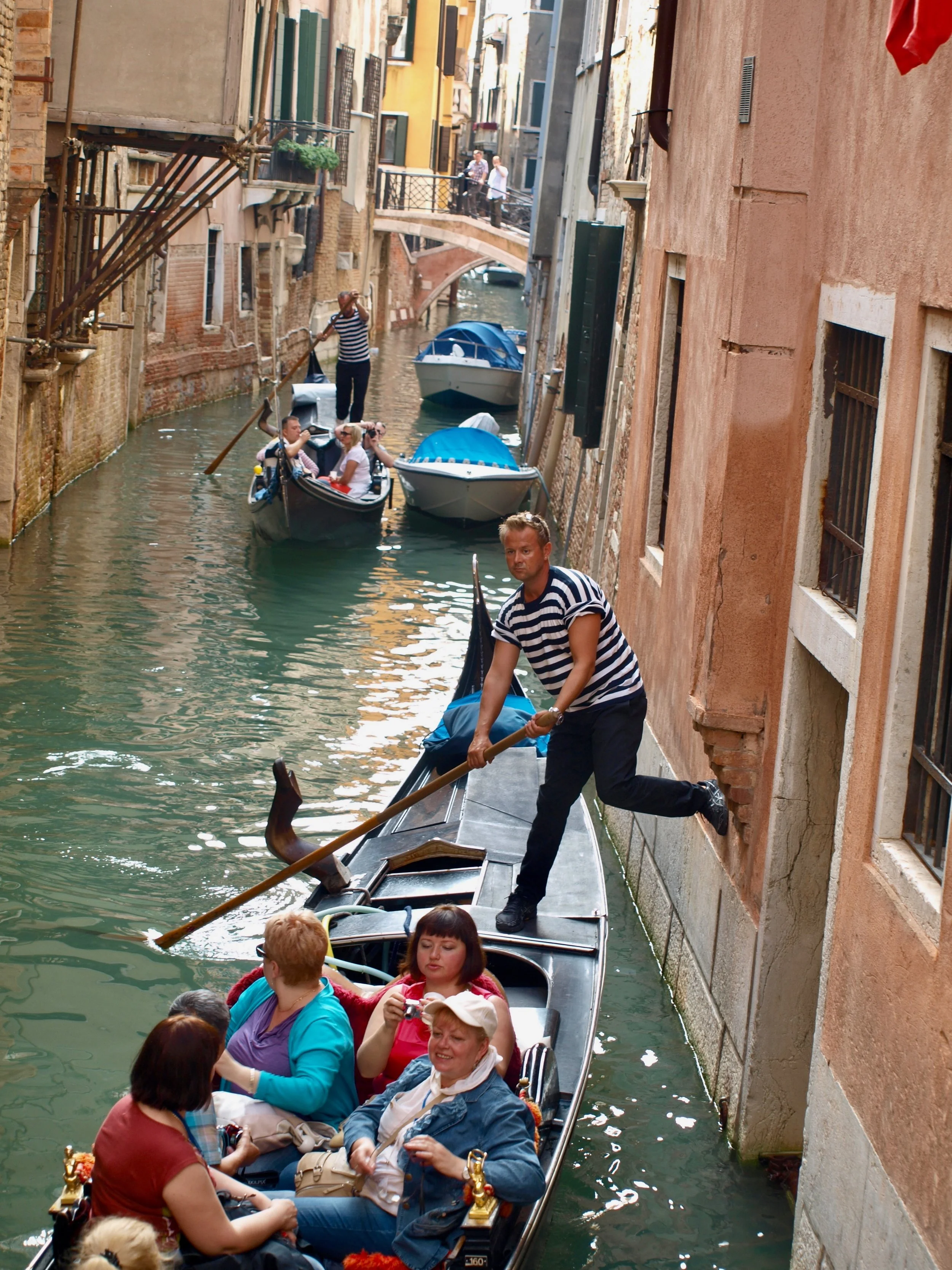 Groupe de touristes dans un bateau sur un canal à Venise, entouré de bâtiments anciens, avec un gondolier en striped shirt en train de pagayer et un autre voyageur avec une caméra.