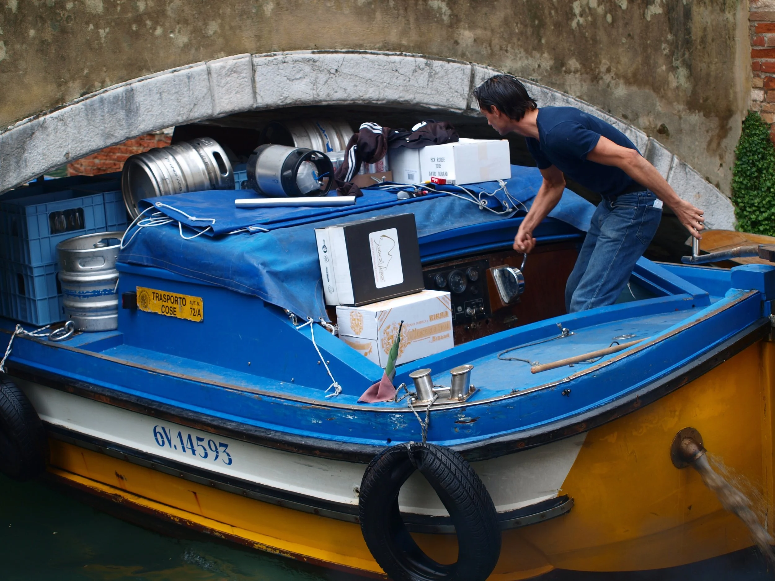 Un bateau bleu avec des fûts en acier et des boîtes sur le pont, stationné sous un pont en pierre, dans un environnement urbain.