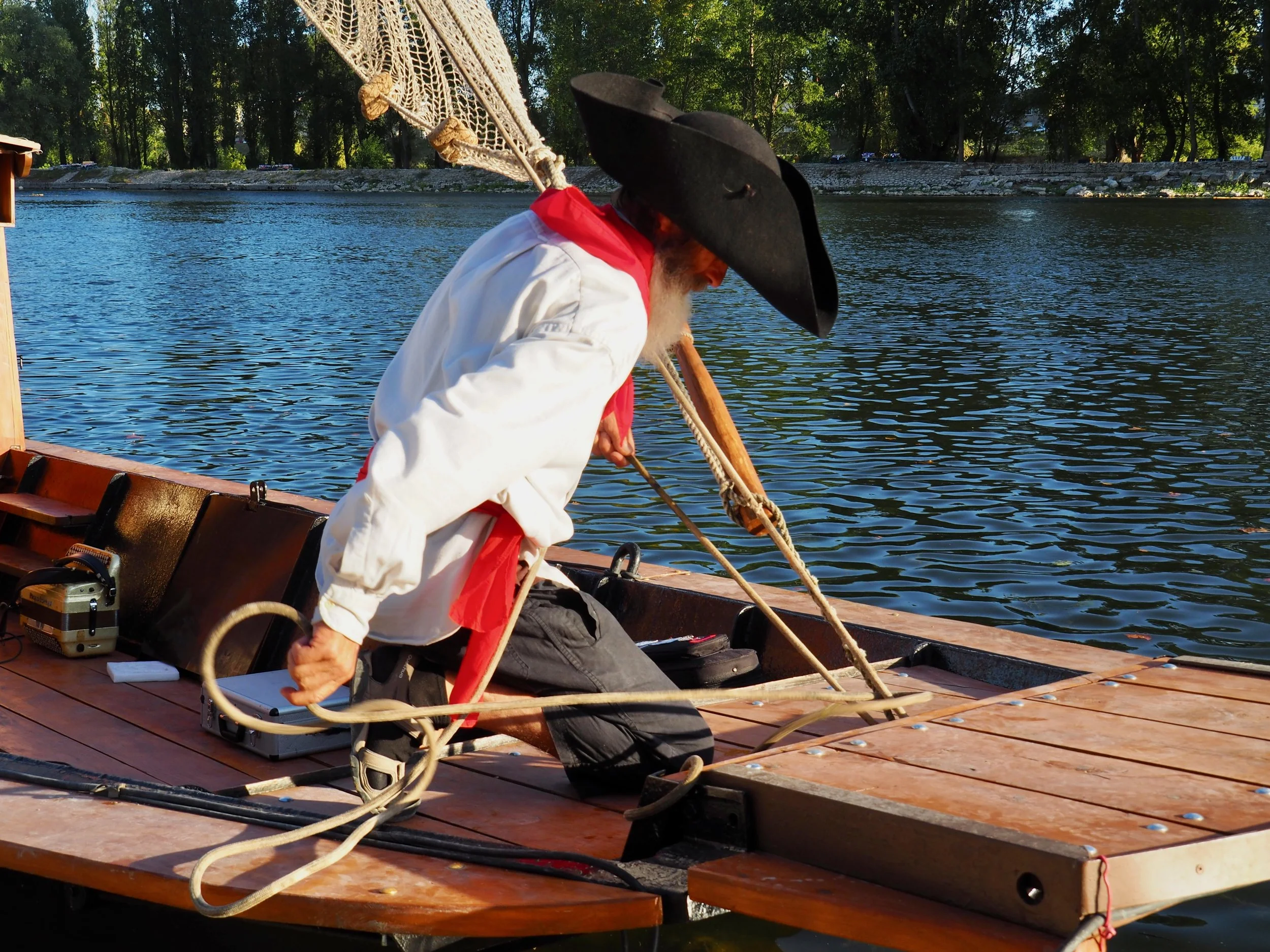 Un homme portant un chapeau noir, une chemise blanche, un foulard rouge, s'agenouille sur un bateau en bois près d'un fleuve, attachant une corde.