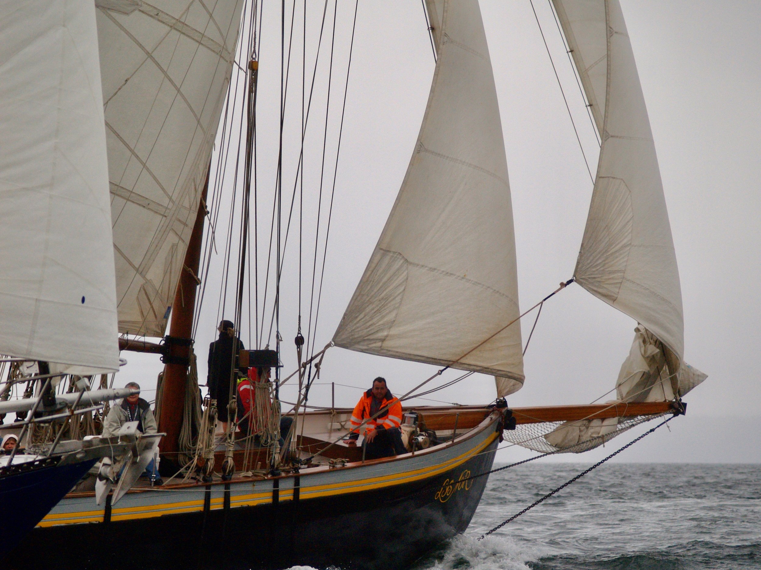 Un voilier avec plusieurs personnes à bord, les voiles sont déployées, et il y a des officiers ou équipiers visibles, dont un portant un manteau orange, en mer par temps nuageux.