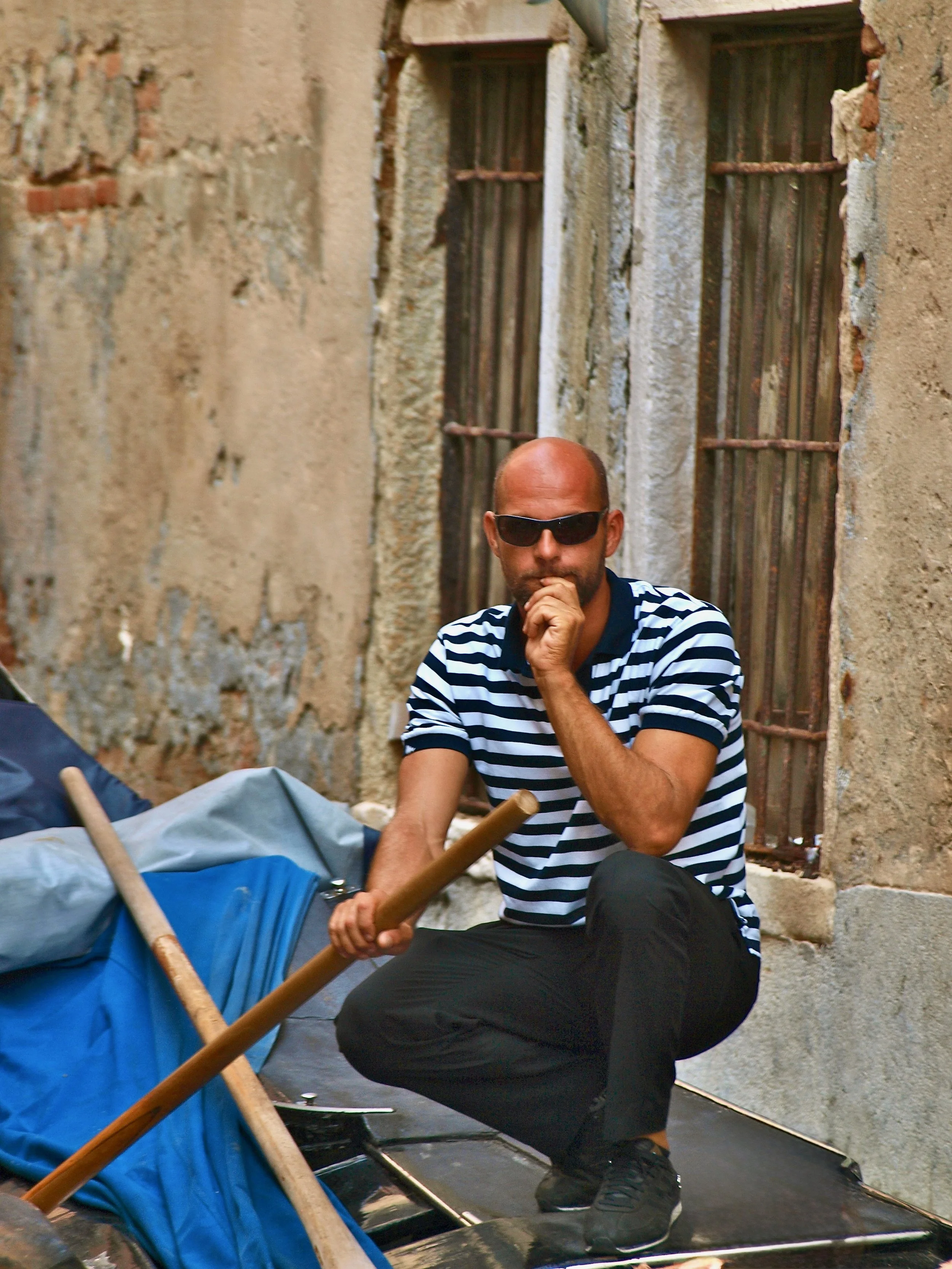 Homme en train de réfléchir, portant des lunettes de soleil et une chemise à rayures, assis contre un mur en pierre avec des fenêtres grillagées.