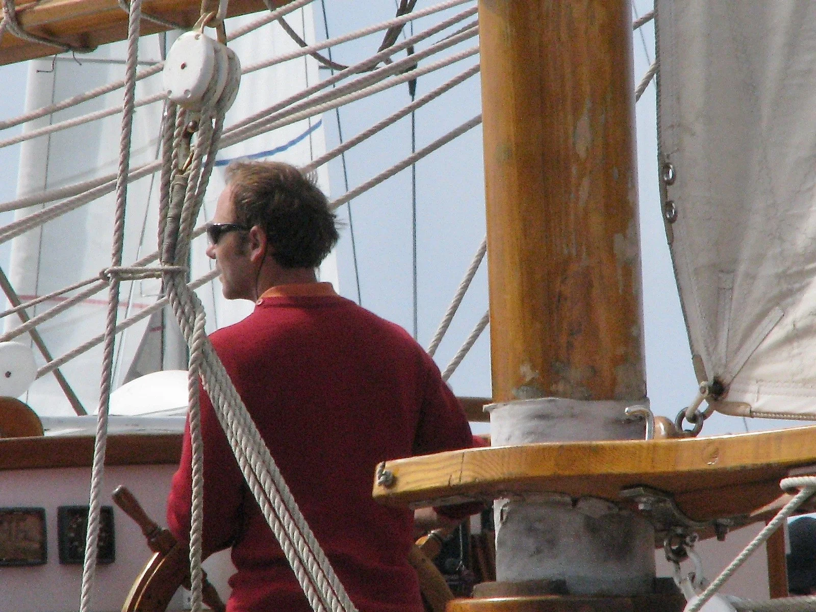 Un homme avec des lunettes de soleil, portant une chemise rouge, se trouve sur un voilier, entouré de cordages et de structures en bois.