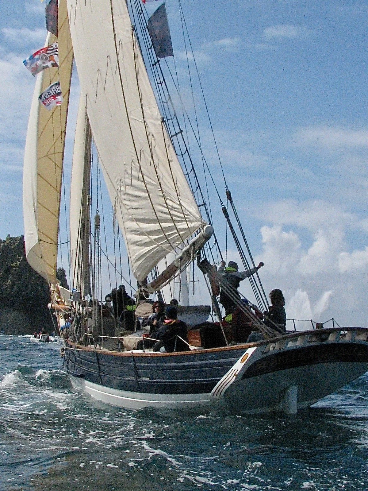 Un voilier navigue en mer avec plusieurs personnes à bord, sous un ciel partiellement nuageux.