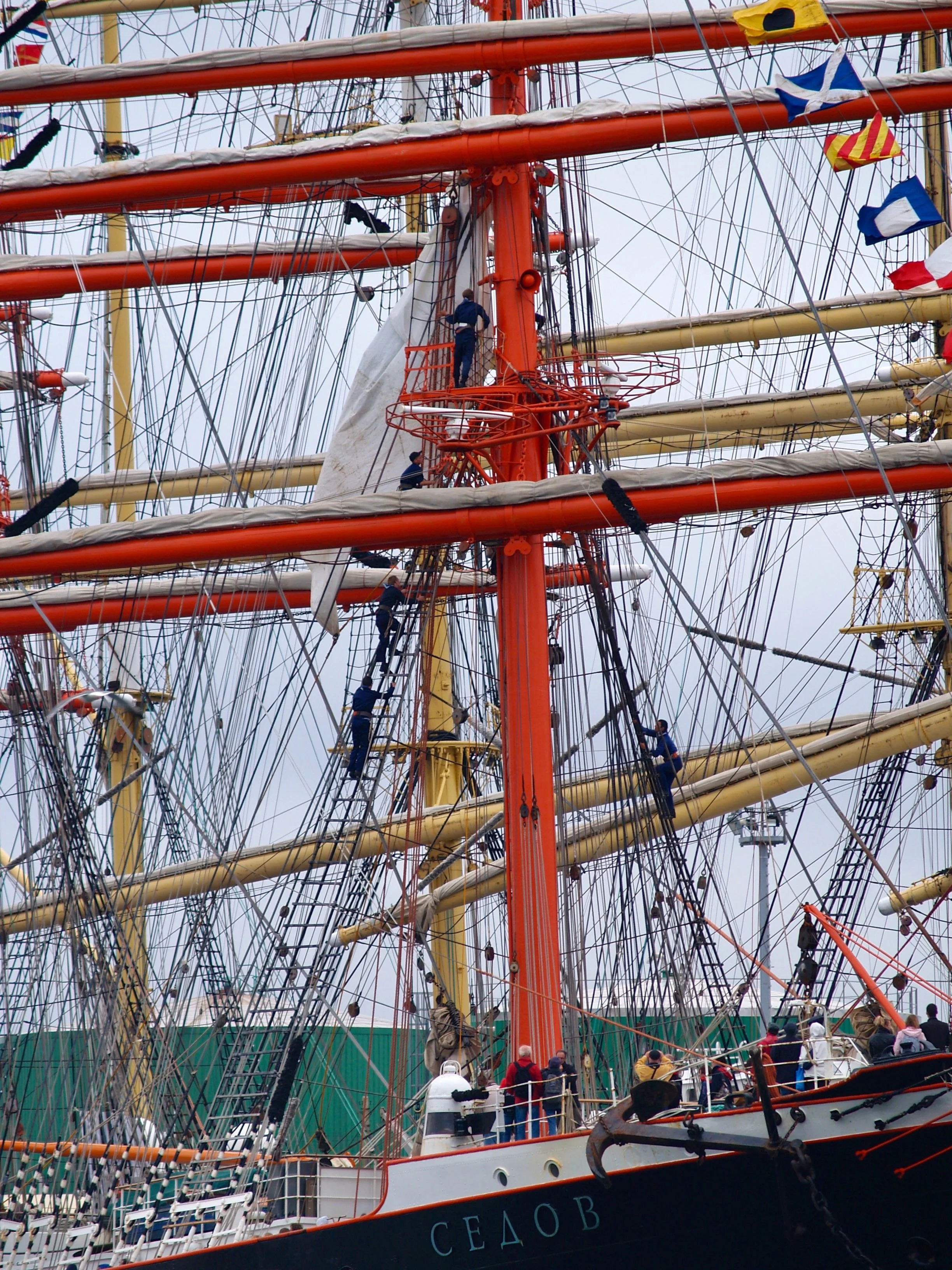 Une grande navigatrice à voiles traditionnel montrée à quai, avec plusieurs personnes sur le pont. Le navire a un grand mât orange, plusieurs voiles et une multitude de cordages fouettés par le vent, avec des drapeaux colorés.