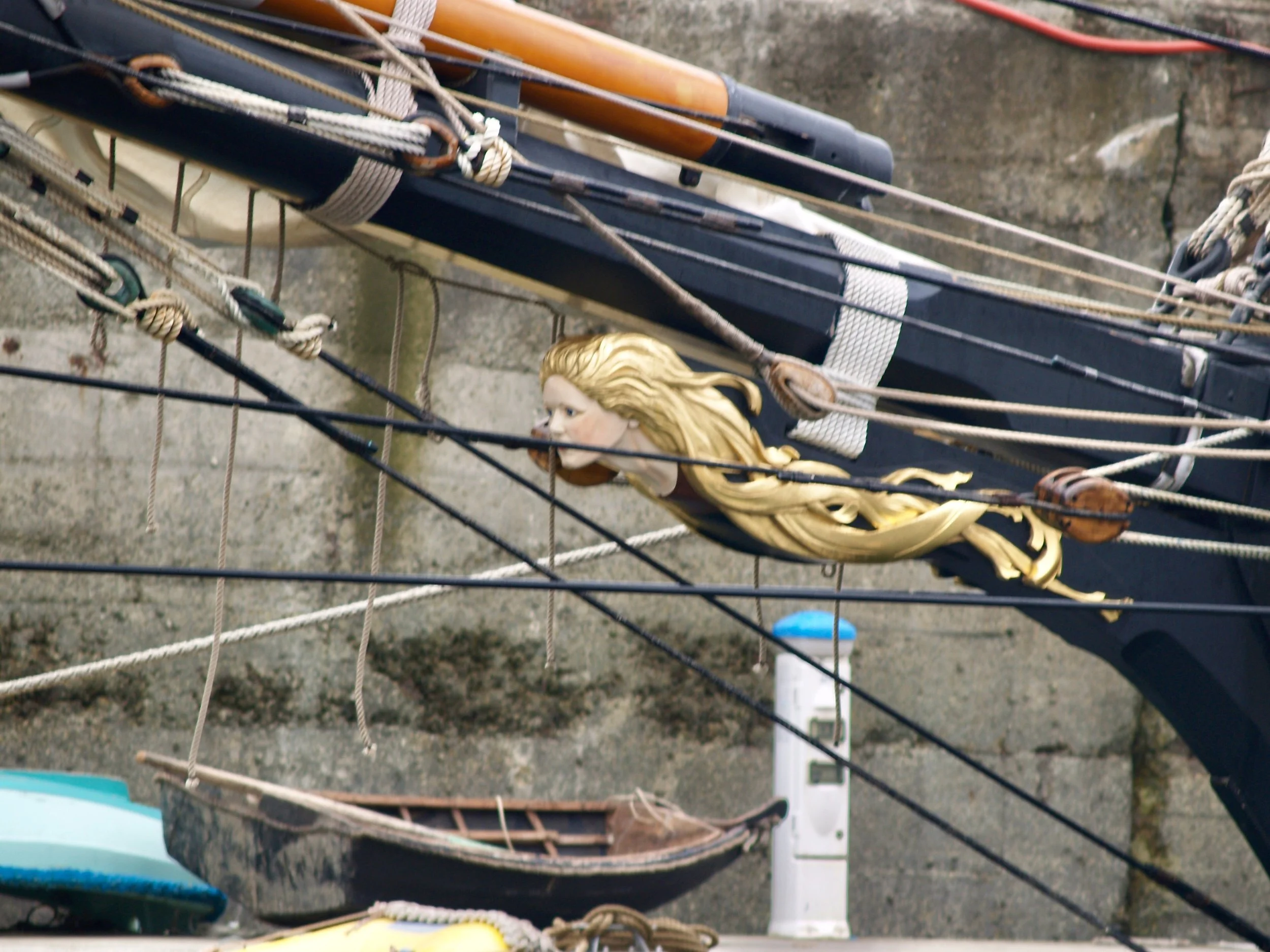 Vue d'une partie d'un bateau à voiles avec des cordages et une figure de proue représentant une femme avec des cheveux longs blondes.