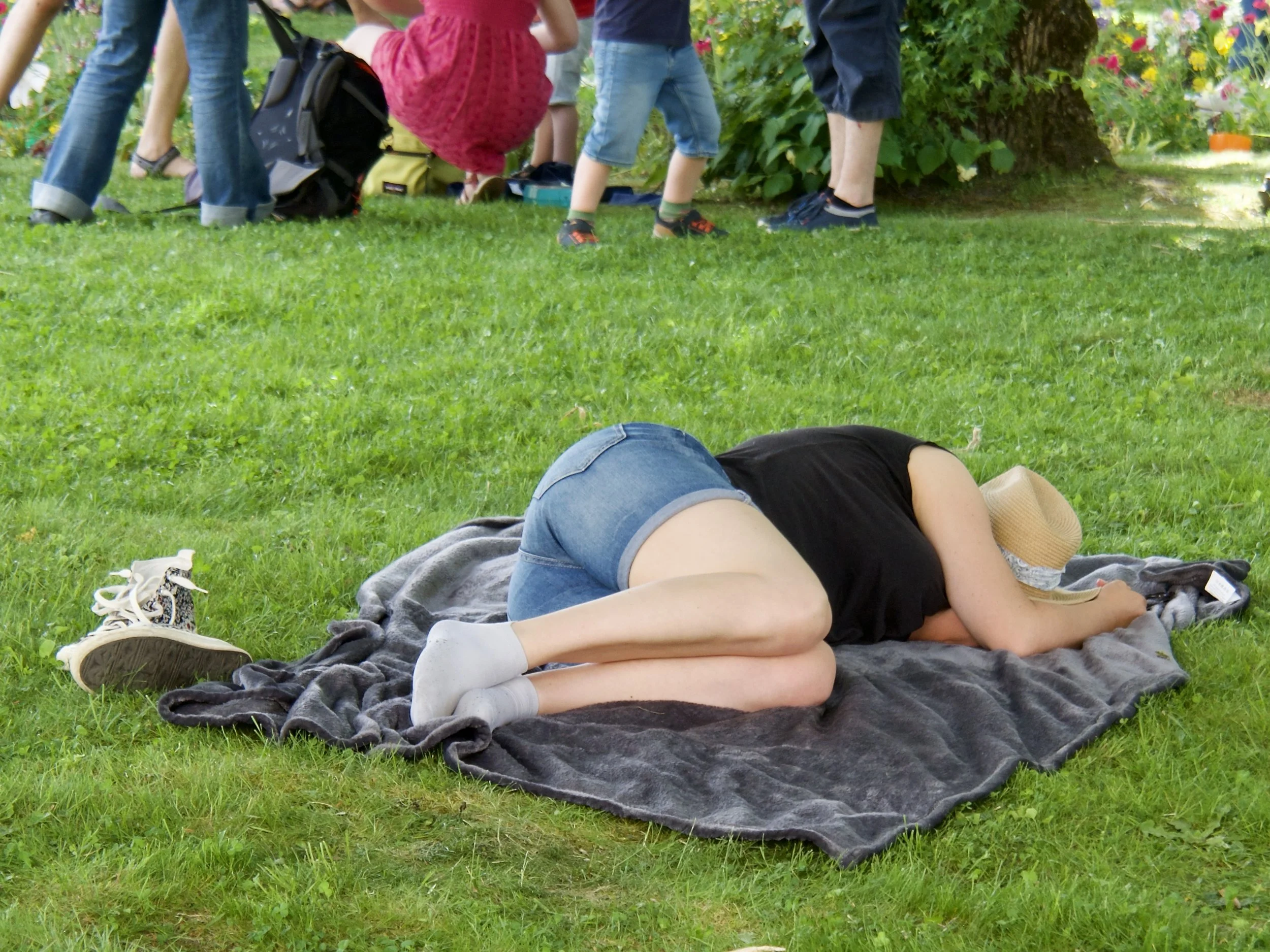 Une personne allongée sur une couverture dans un parc, vue du dessous, avec un groupe d'enfants en arrière-plan près d'un arbre.