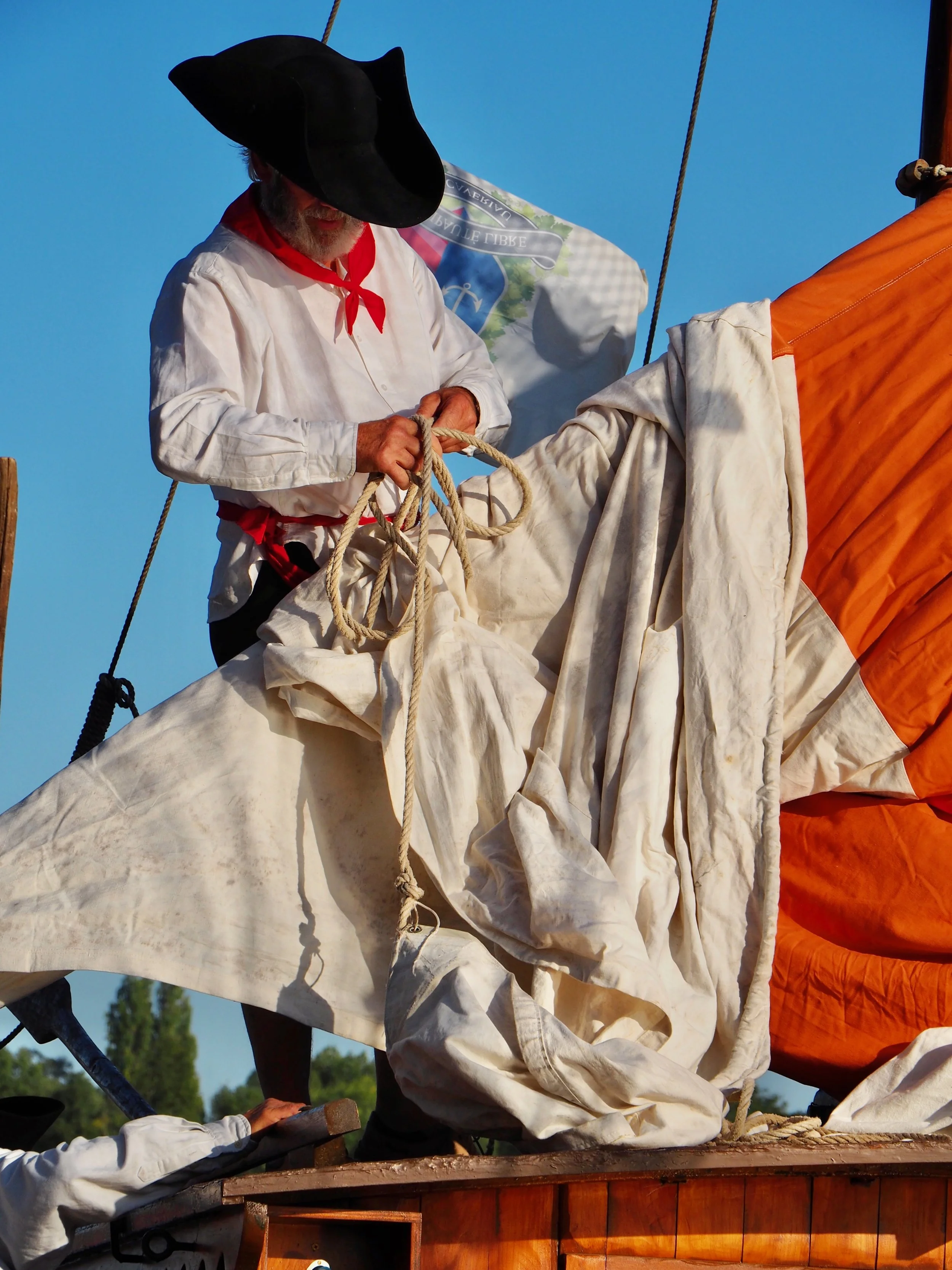 Un homme vêtu d'une chemise blanche, portant une large casquette noire et une étole rouge, est en train de plier des cordages sur une embarcation. Derrière lui, un drapeau blanc avec un logo et un fond de ciel bleu clair.