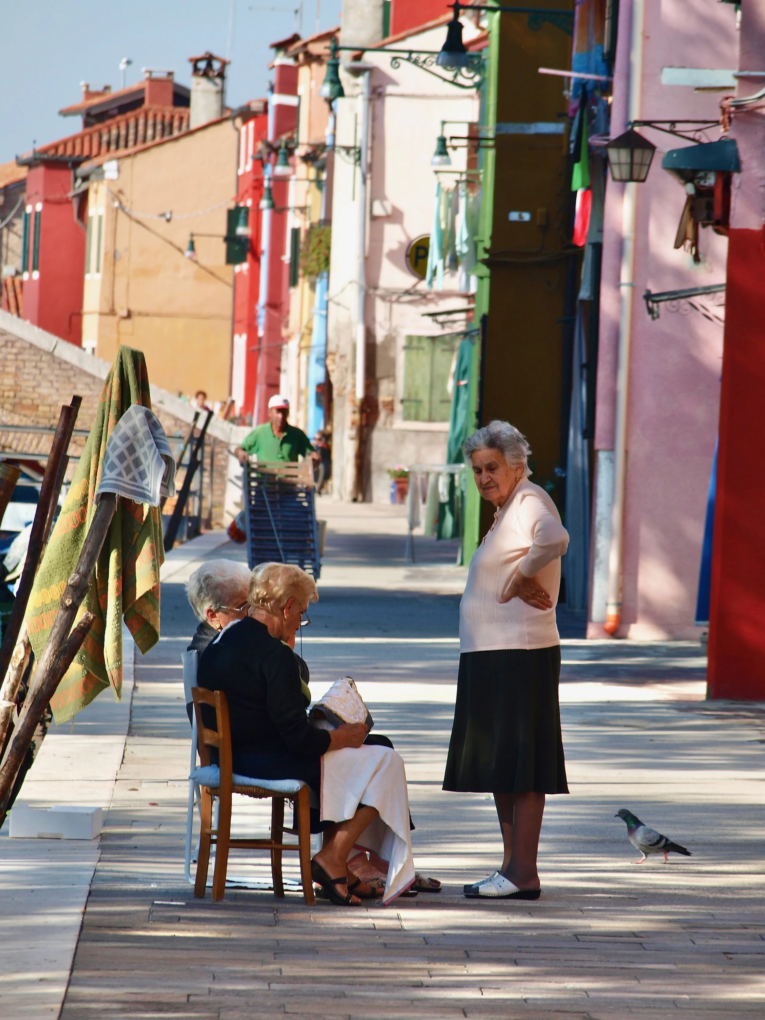 Une vieille femme assise sur une chaise en bois, une femme plus jeune se tenant debout à côté, dans une rue en plein air avec des maisons colorées, et un pigeon au sol devant elles.