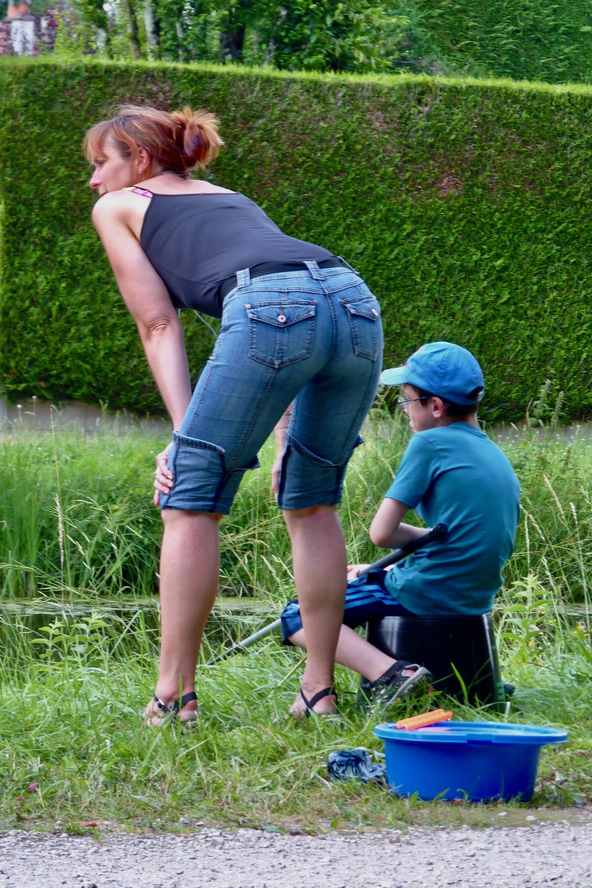 Une femme et un garçon près d'une rivière, la femme se penche en avant, le garçon assis sur un seau, dans un environnement naturel avec des plantes et un arbre en arrière-plan.