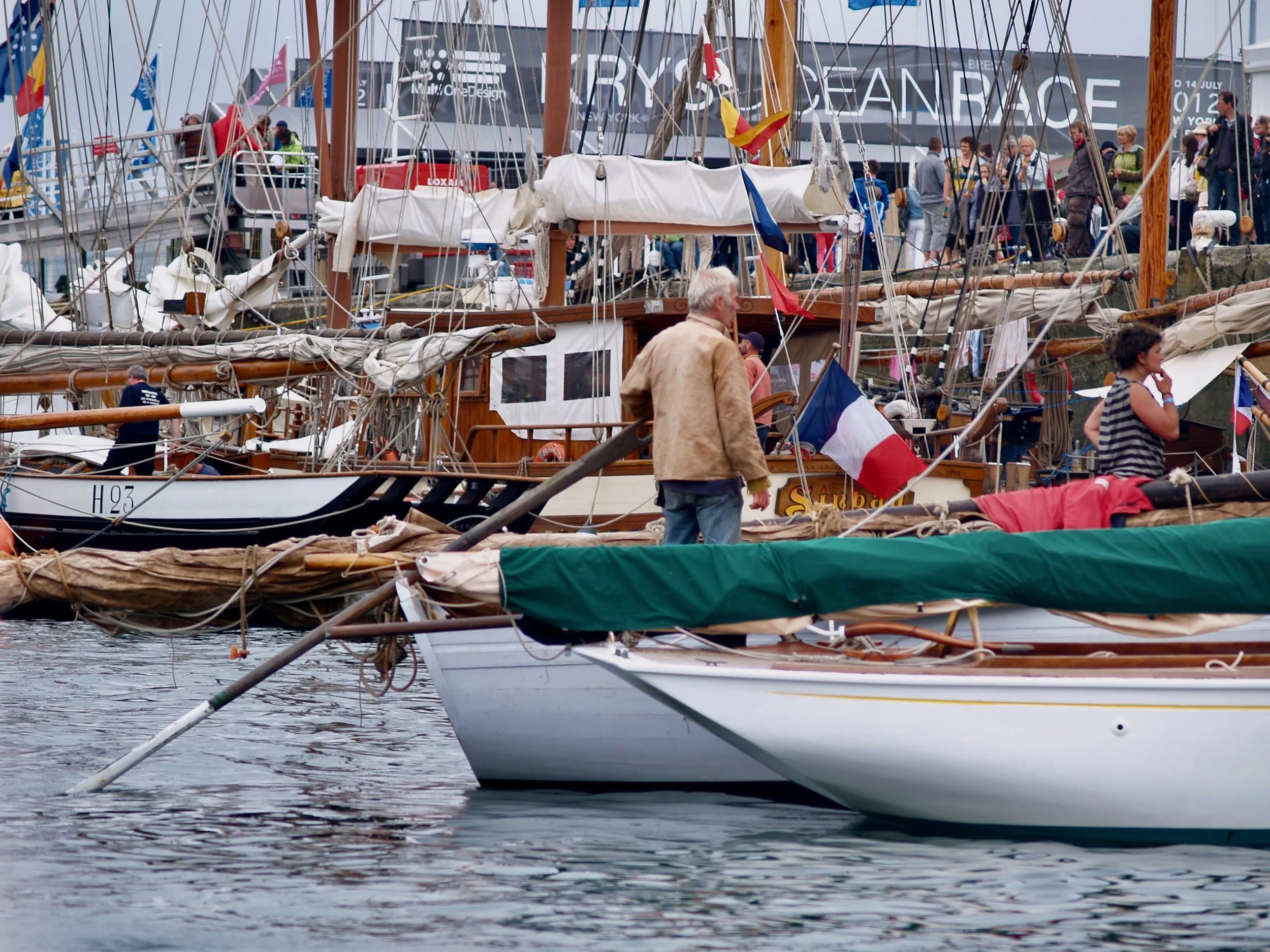 Un bateau à voile en bois avec des voiles en tissu blanc, des drapeaux français et d'autres couleurs, amarré dans un port lors d'un événement nautique. Plusieurs personnes sont visibles sur le bateau et le quai, certaines regardant le bateau, d'autre