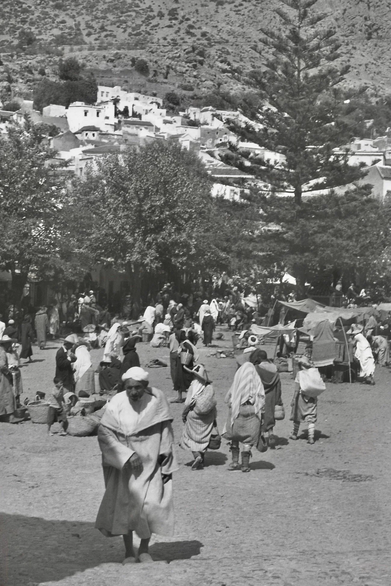 Une scène de marché animée en plein air avec de nombreuses personnes portant des vêtements traditionnels, des chapeaux et des paniers, sous des arbres, avec des maisons blanches sur une colline en arrière-plan.