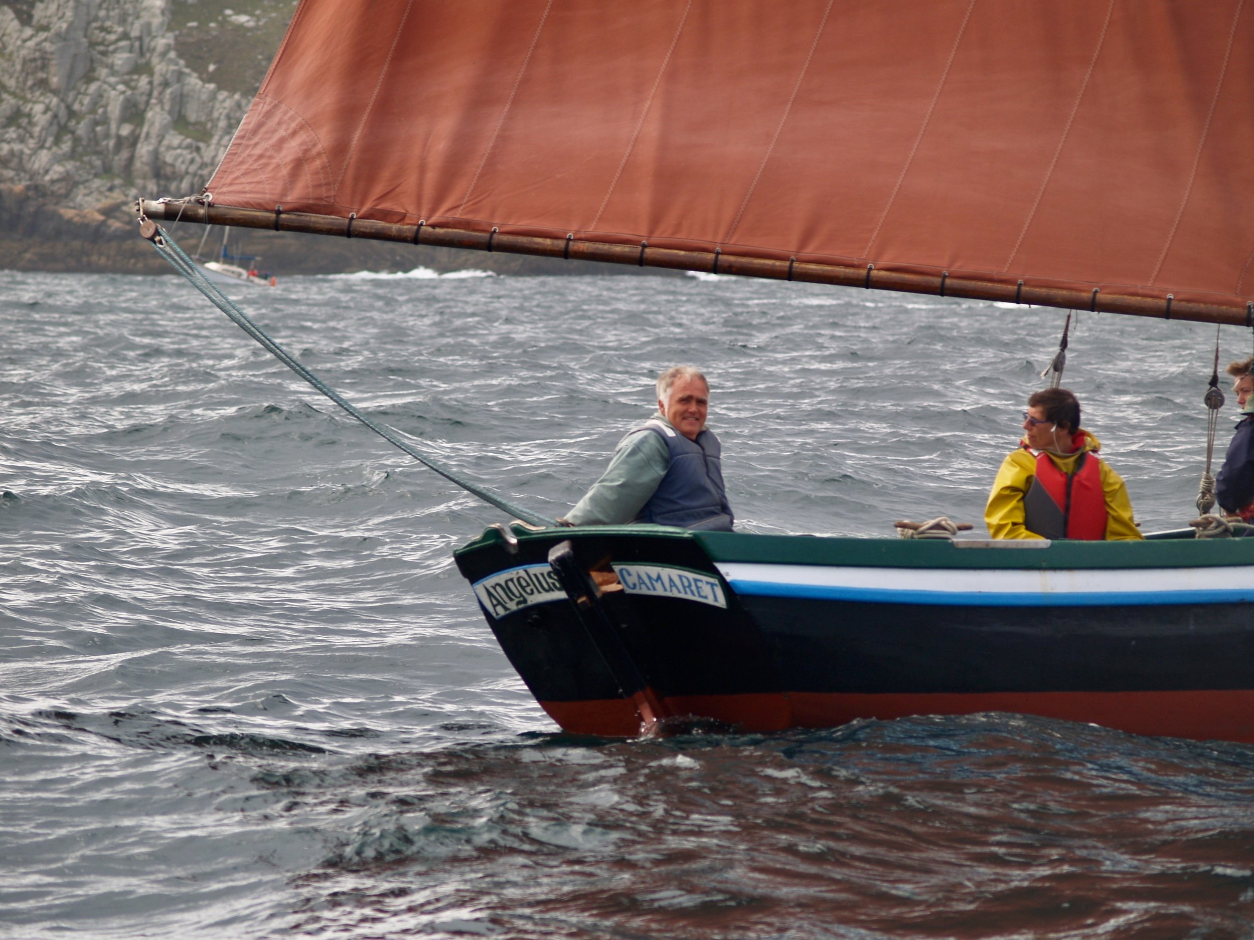 Trois personnes naviguent en voilier sur la mer, avec des rochers en arrière-plan et un grand voilier dont la voile est visible au-dessus d'eux.