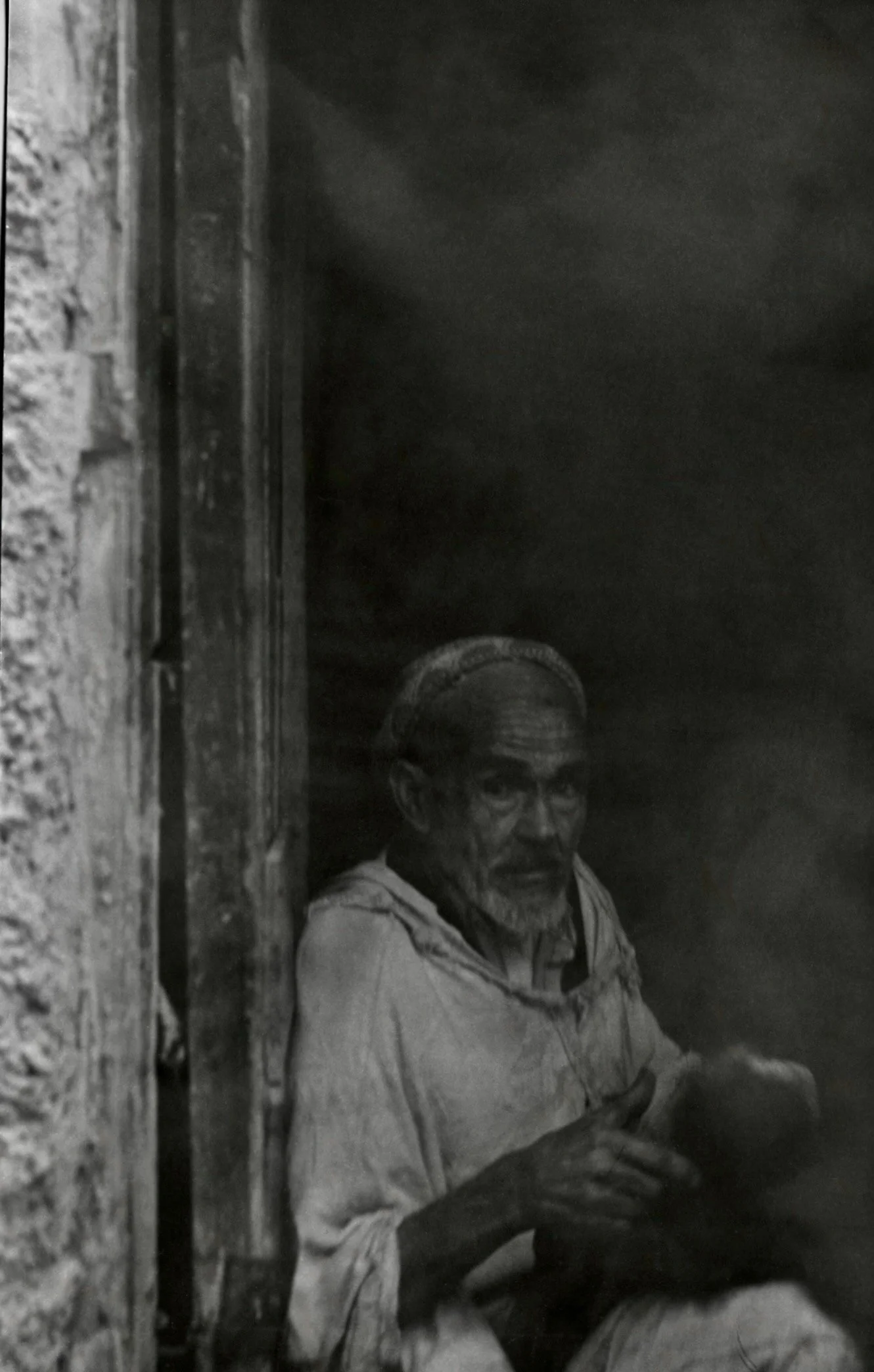 Un homme âgé à lunettes, assis dans un espace sombre, portant une veste et un t-shirt, avec une expression sérieuse.