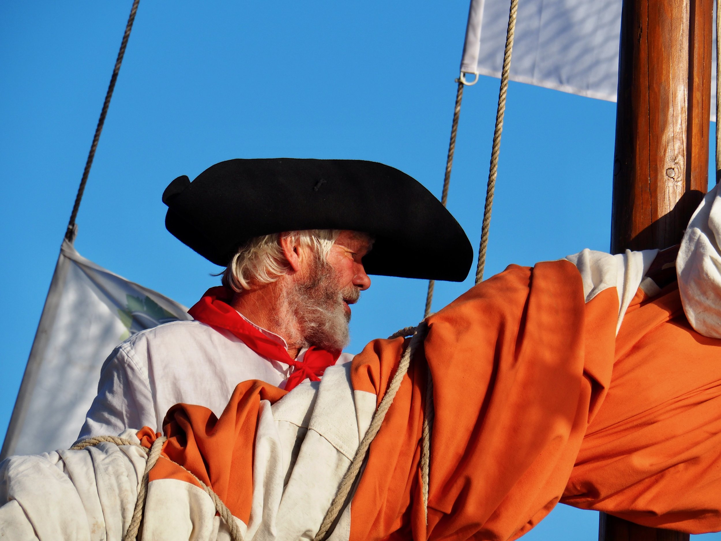 Un homme âgé portant un chapeau noir et une veste orange, il tient une corde et semble être à bord d'un voilier contre un ciel bleu clair.