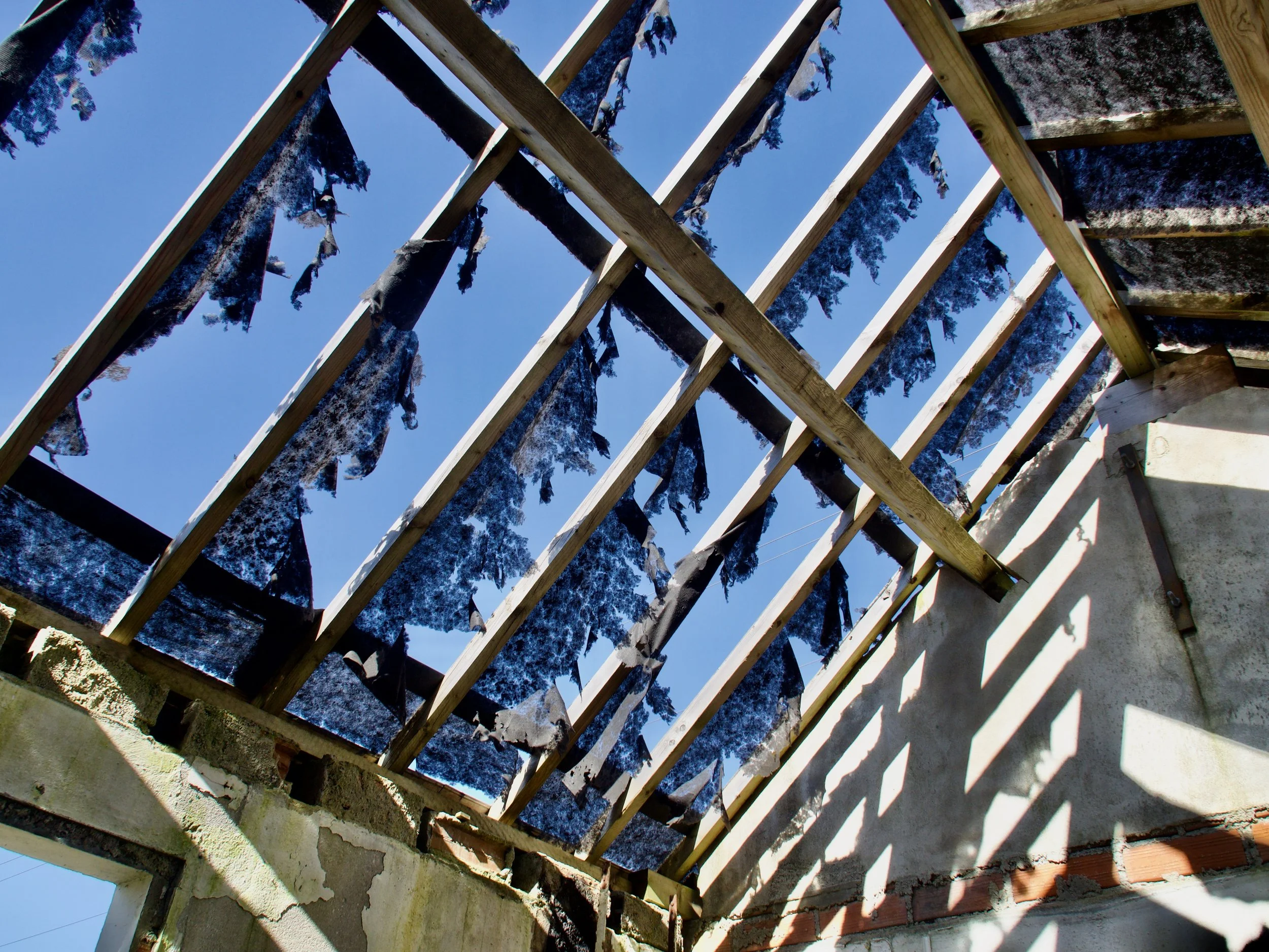 Toit d'une structure en construction avec des planches de bois et des morceaux de tissu noir suspendus, vue en contre-plongée contre un ciel bleu.