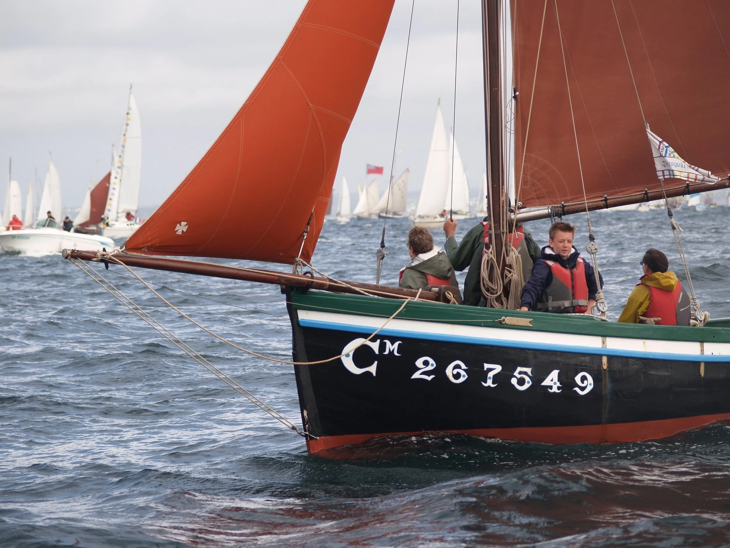 Des voiliers avec des voiles orange et blanches participent à une régate sur la mer, avec d'autres bateaux en arrière-plan, par un jour nuageux.