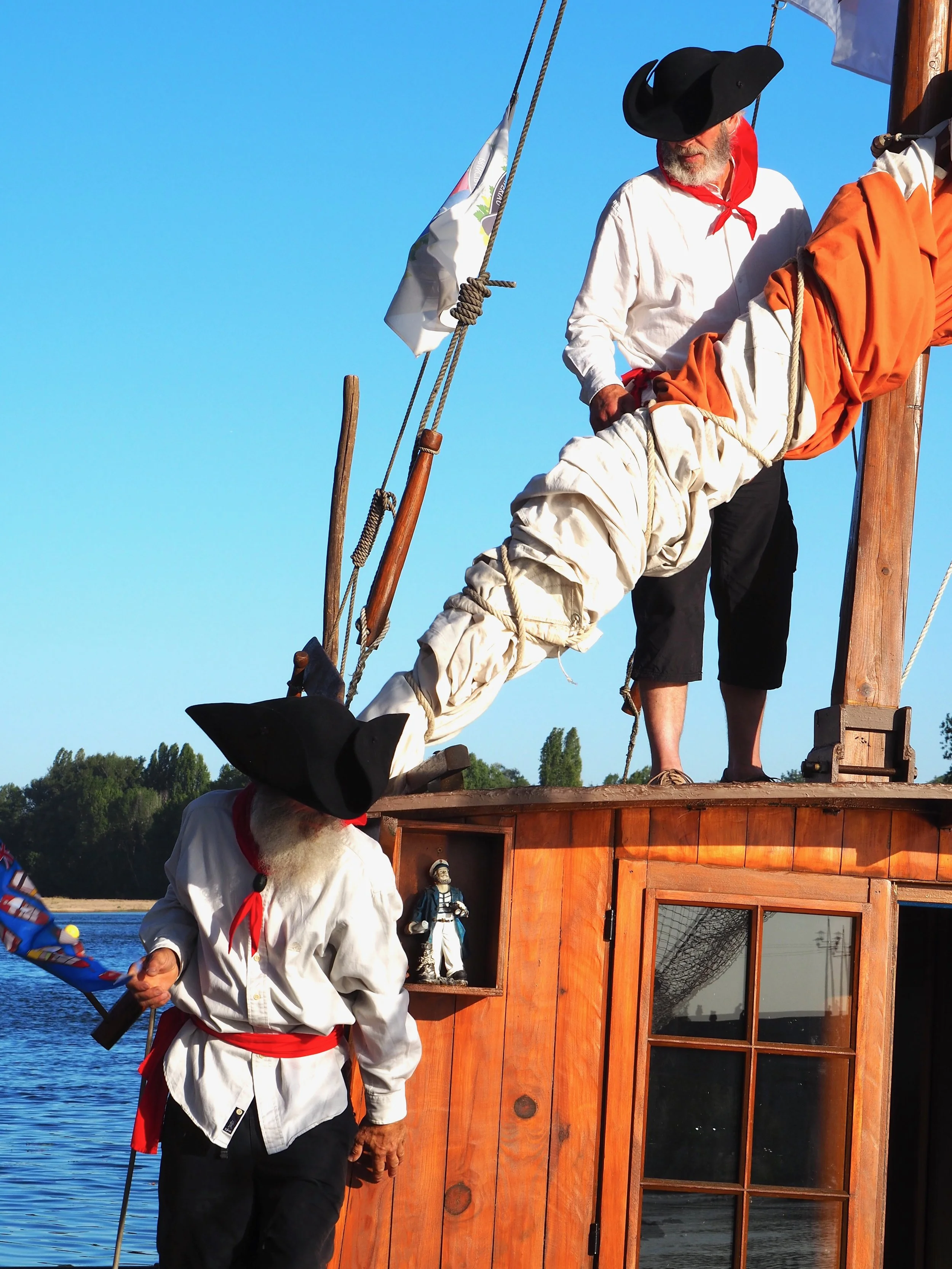 Personne portant un costume traditionnel de marin, à côté d’un bateau en bois, avec un drapeau, sur un lac ou une rivière, sous un ciel bleu.