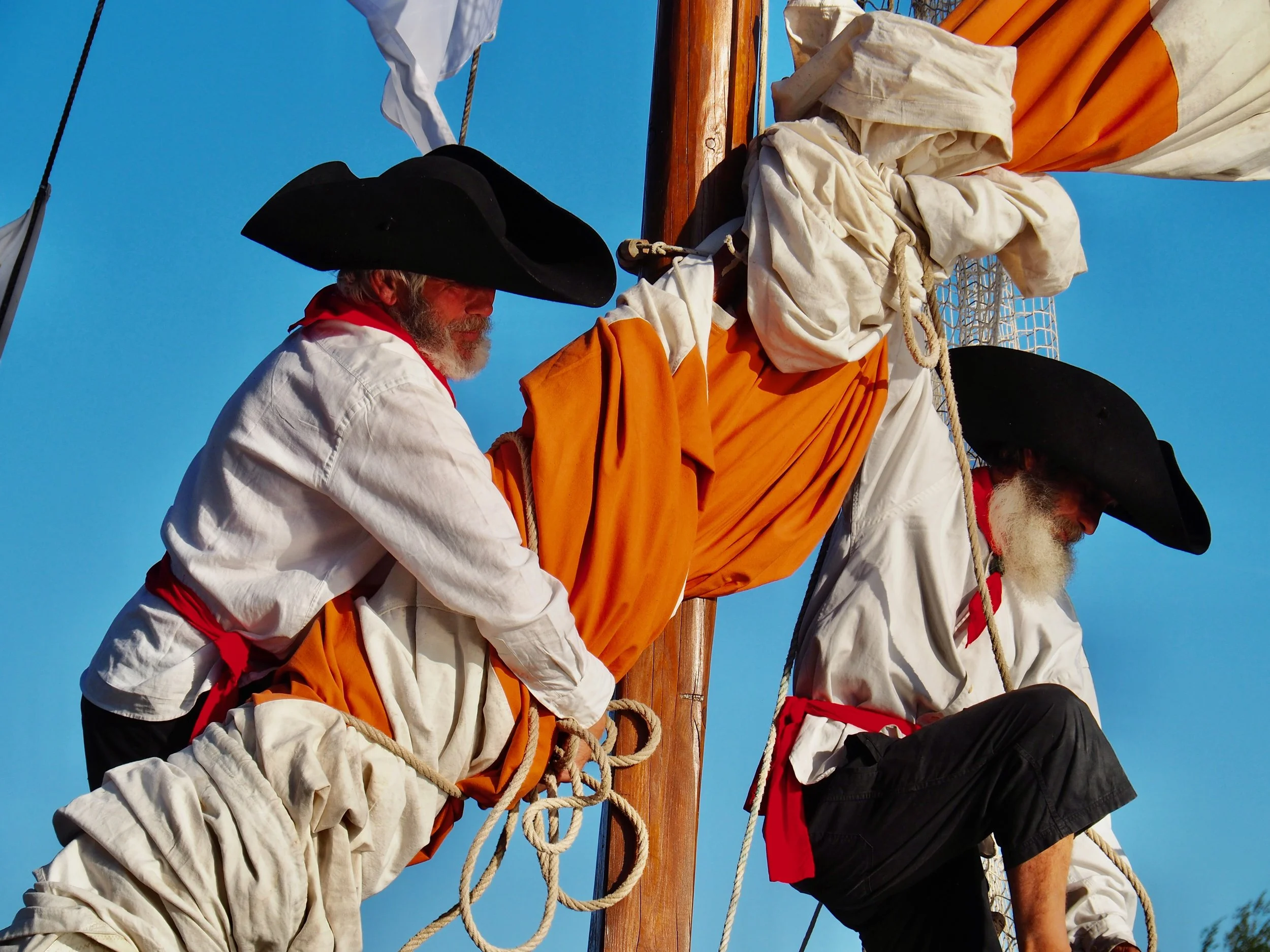 Trois hommes en costumes historiques, portant des chapeaux noirs, fabriquant une croix sur un mât en bois lors d'une cérémonie en plein air sous un ciel bleu.