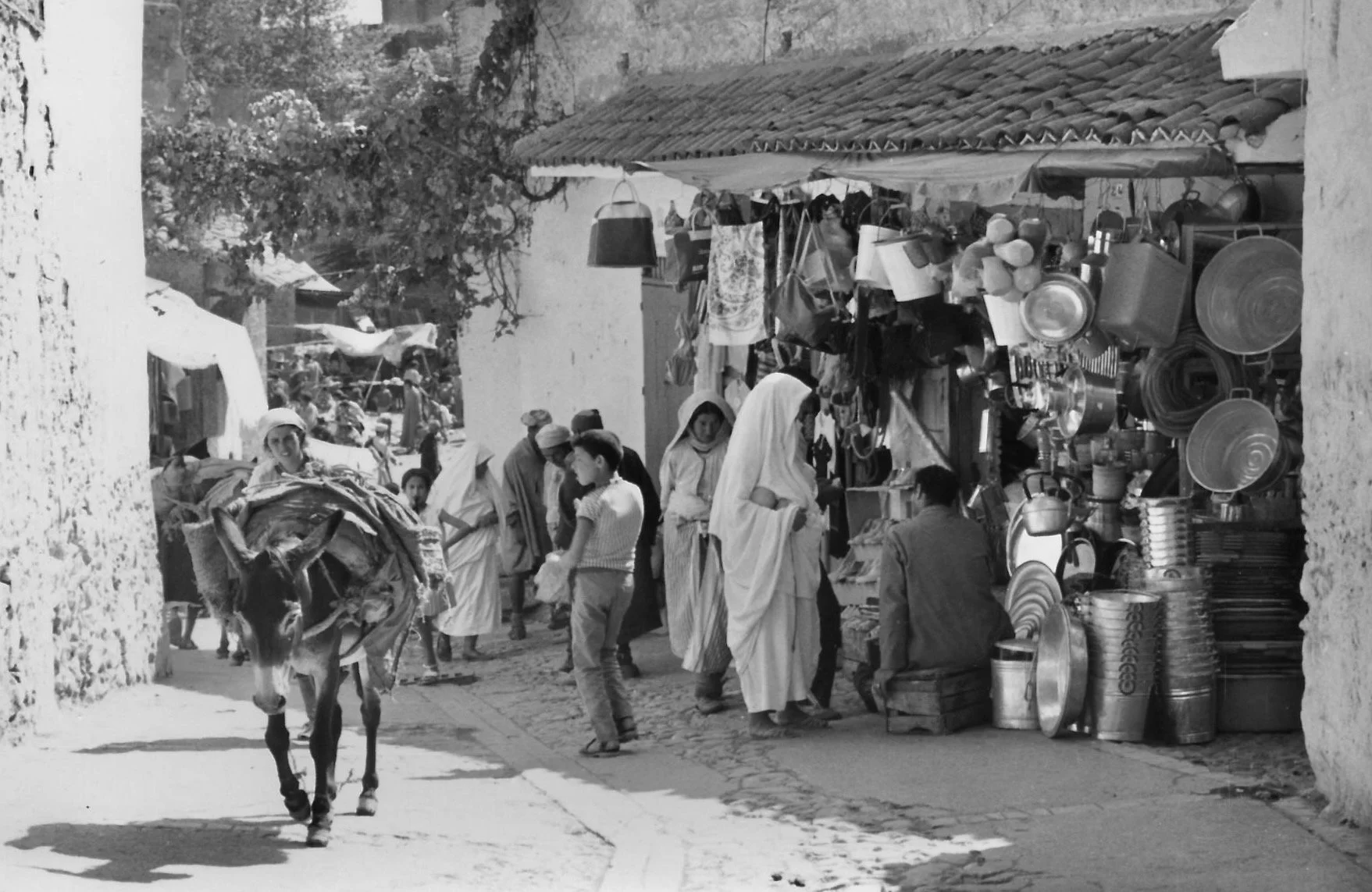 Une rue animée avec des personnes marchant, un vendeur avec un âne chargé de marchandises, et un étalage de vaisselle en métal et autres articles. Des femmes en vêtements traditionnels et enfant en discussion. Scène de marché en plein air.