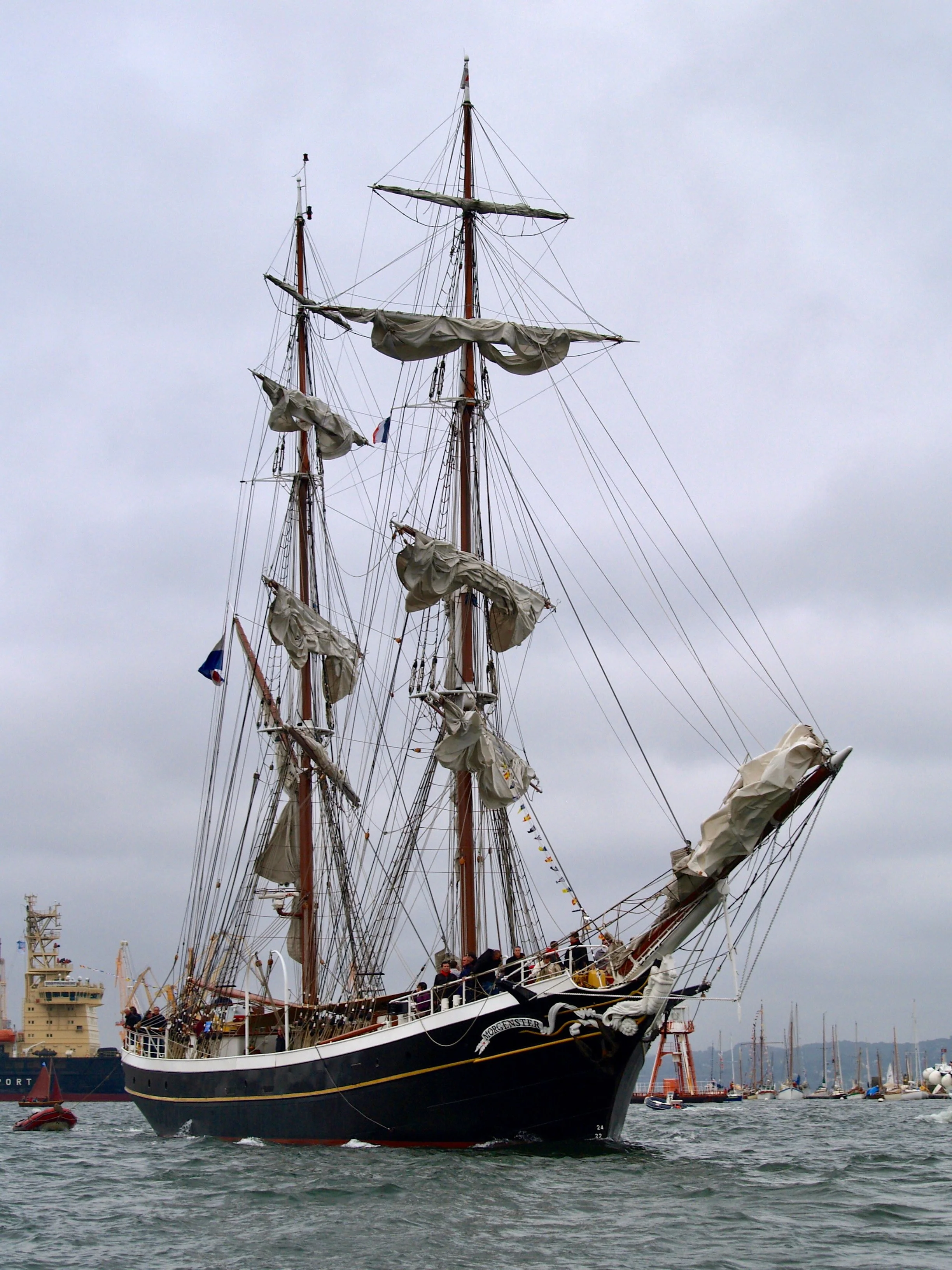 Un grand navire à voile traditionnel avec plusieurs mats déployés, des voiles enroulées, navigue sur l'eau, avec un port et d'autres voiliers en arrière-plan, un ciel nuageux.