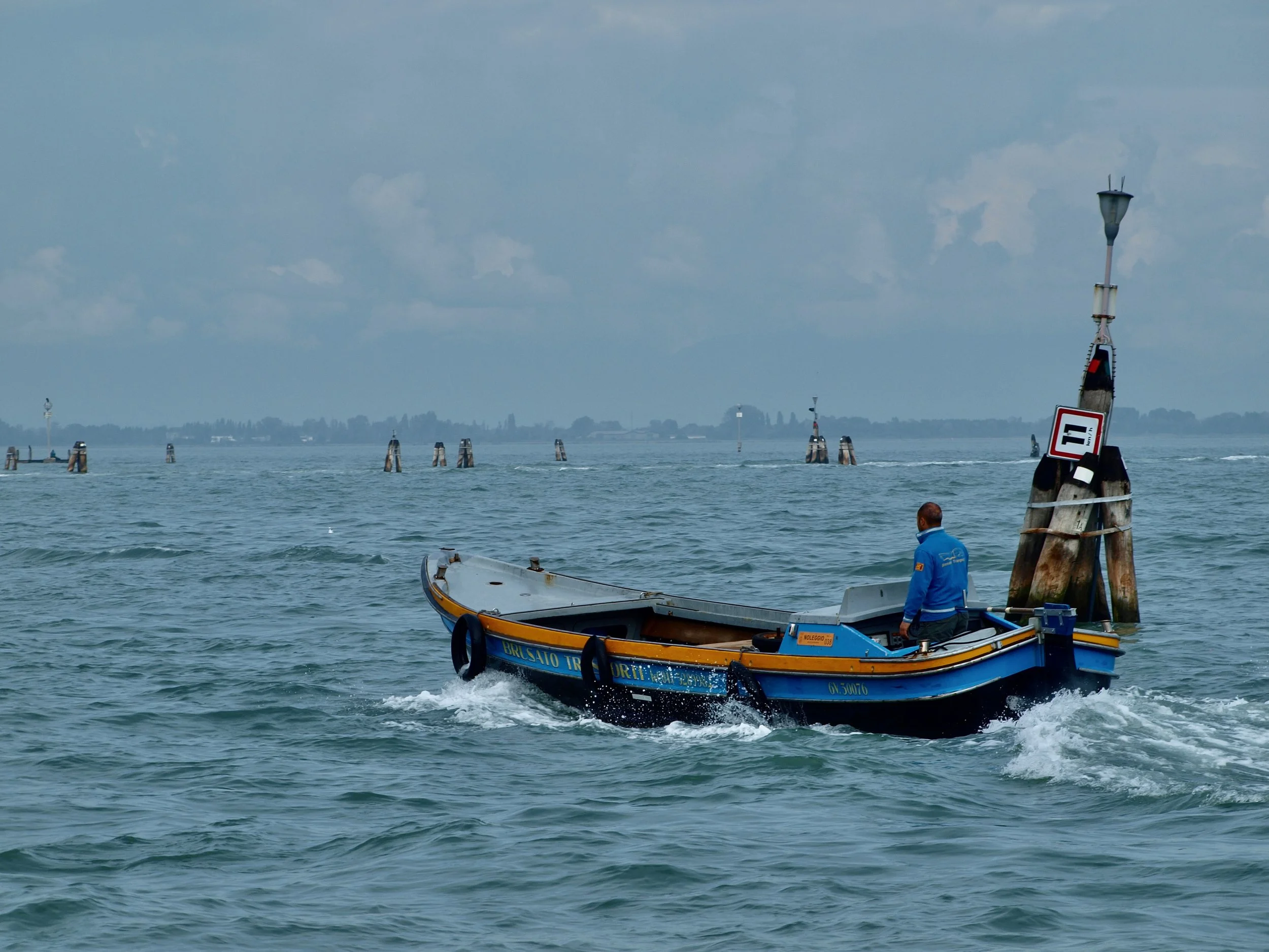 Un bateau de pêche en mer avec un homme en veste bleue à bord, entouré de bouées, et un lampadaire flottant dans l'eau avec des panneaux et des balises en arrière-plan.