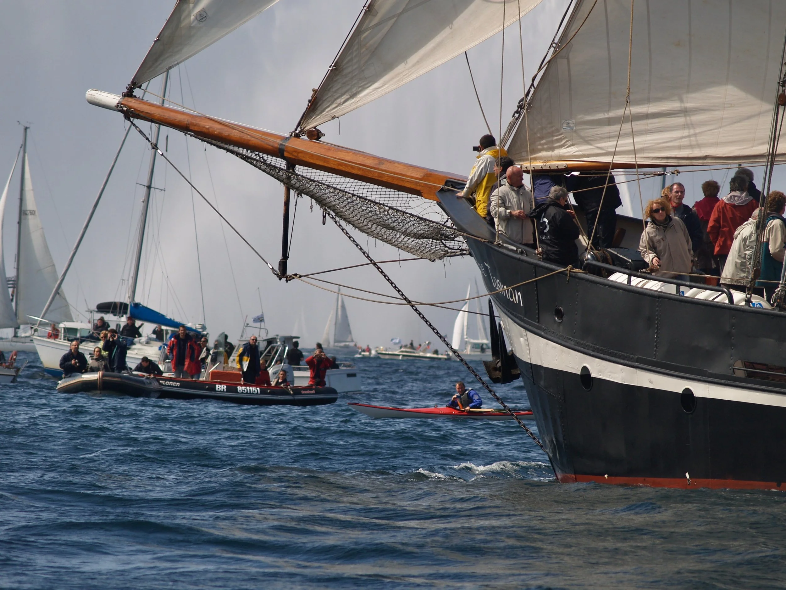 Un voilier navigue en mer avec plusieurs personnes à bord, entouré d'autres bateaux de plaisance.