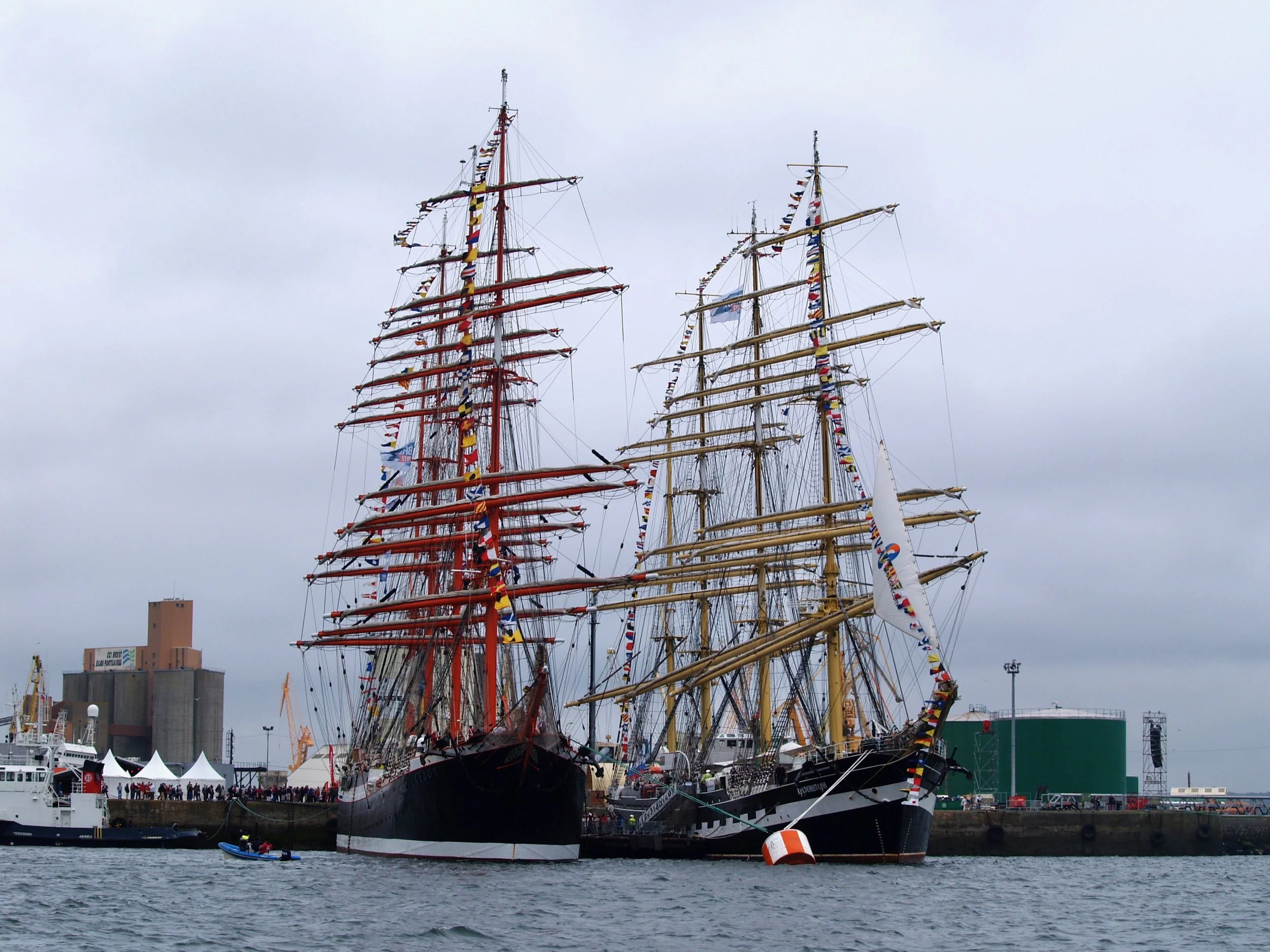 Deux grands voiliers avec des mâts colorés et des guirlandes de drapeaux flottant dans un port, avec des bâtiments et des bateaux plus petits en arrière-plan.