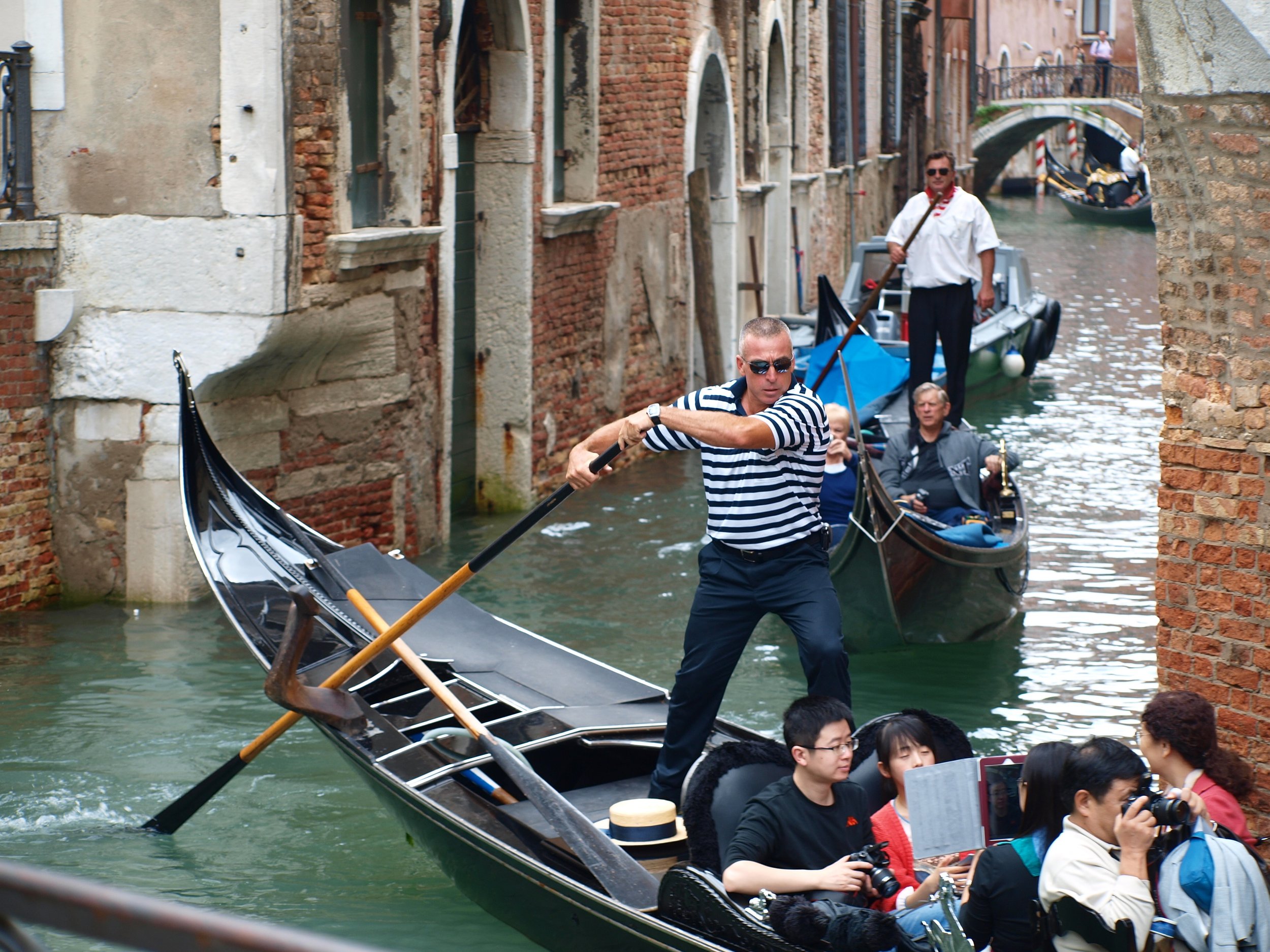 Gondoleurs naviguant une gondole sur un canal à Venise, avec des bâtiments anciens en pierre et briques en arrière-plan.