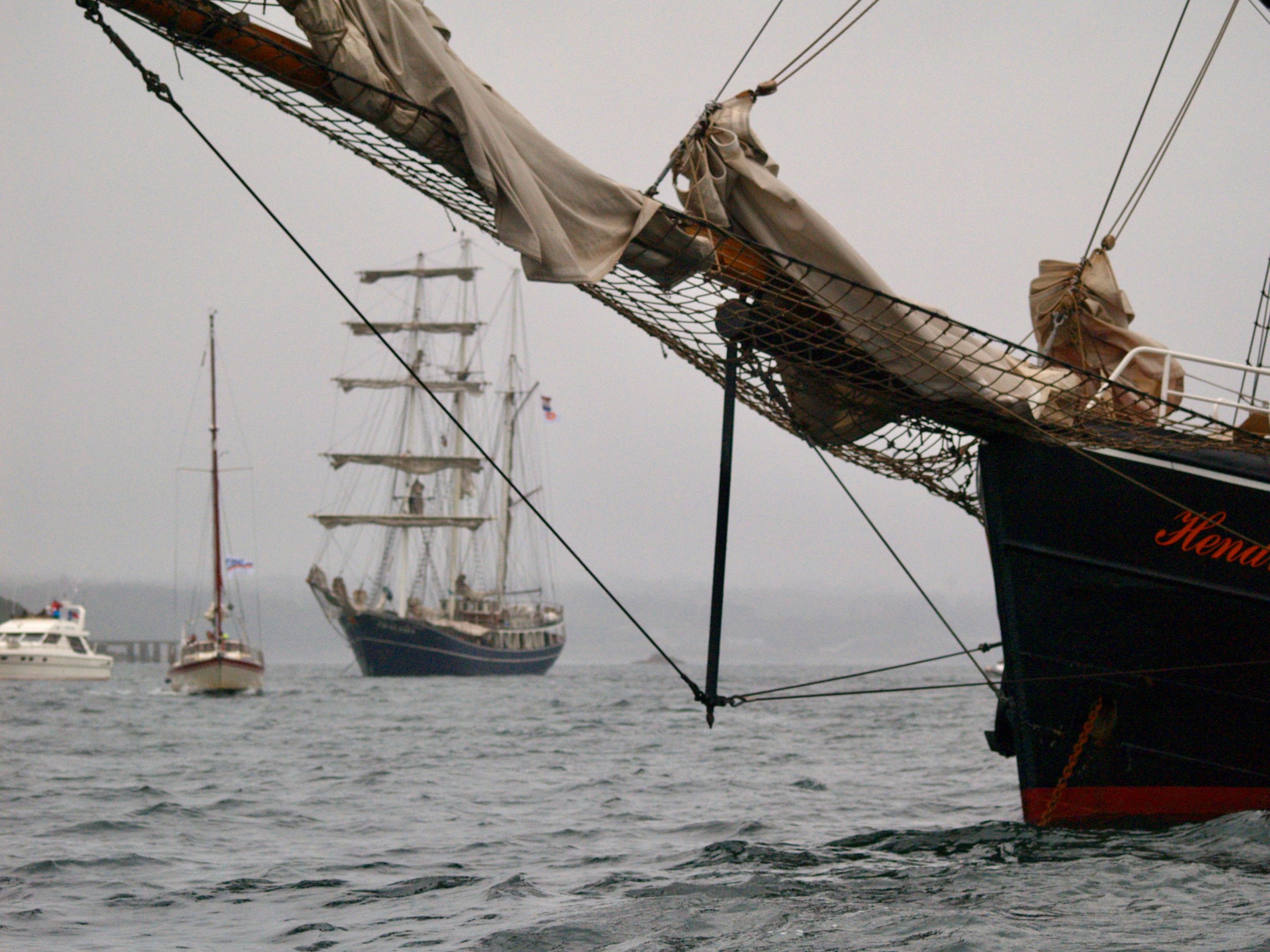 Plusieurs grands voiliers amarrés en mer, avec un focus sur la proue du voilier en premier plan, montrant les voiles et la coque noire avec une inscription rouge. Le ciel est gris et la mer calme.