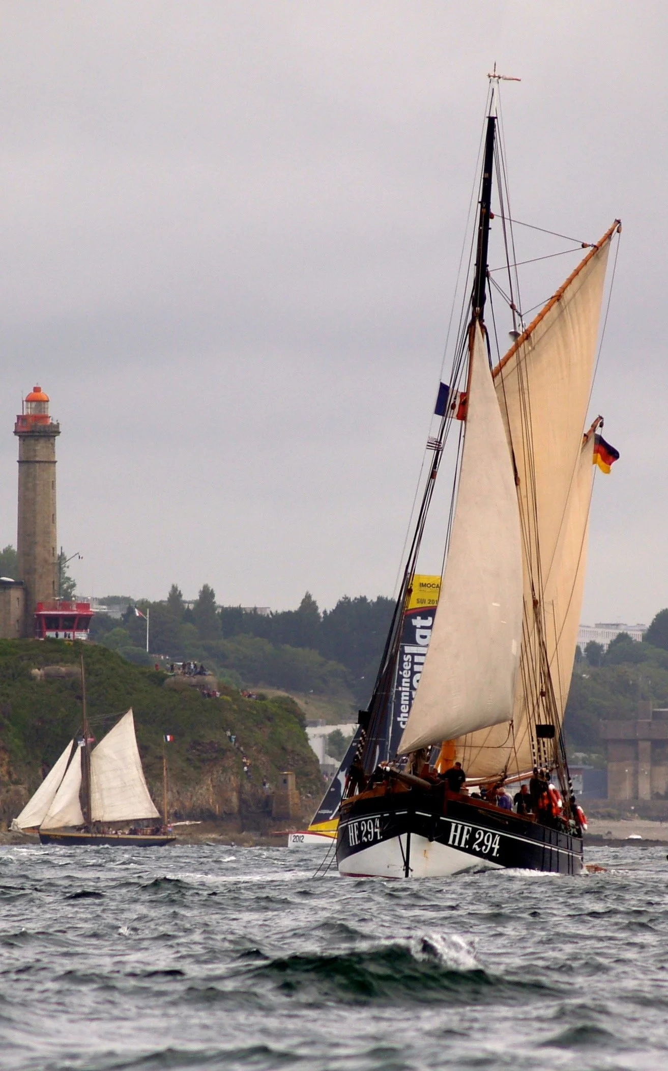 Un voilier en mer avec ses voiles déployées et un phare sur la côte en arrière-plan.
