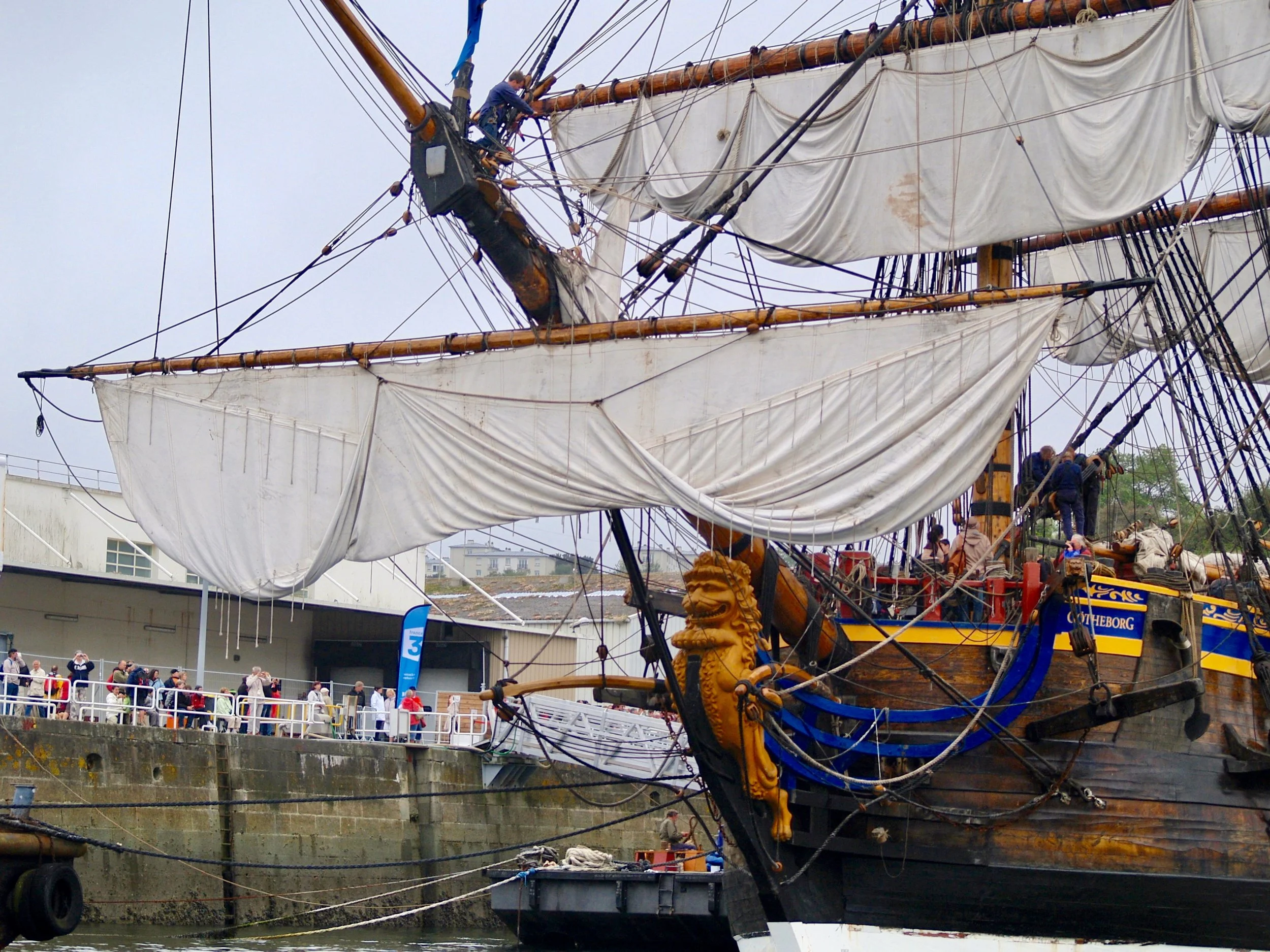 Un grand voilier en bois avec plusieurs mâts et voiles, exposé lors d'une manifestation ou d'une visite guidée, avec des personnes qui montent et descendent à bord.