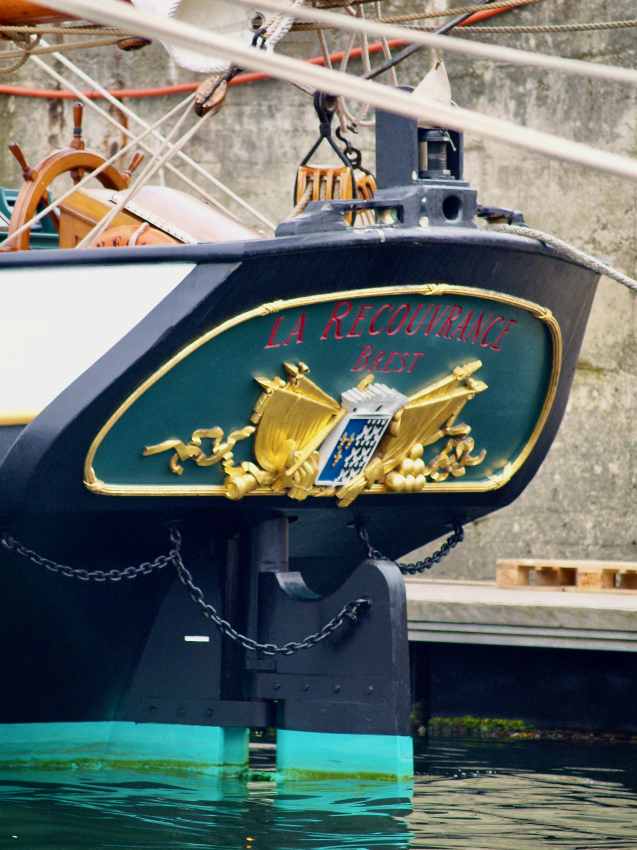 Vue rapprochée de la proue d'un bateau nommé La Recouvrance, avec des décorations dorées, un emblème au centre et des cordages, amarré au quai avec l'eau en dessous.