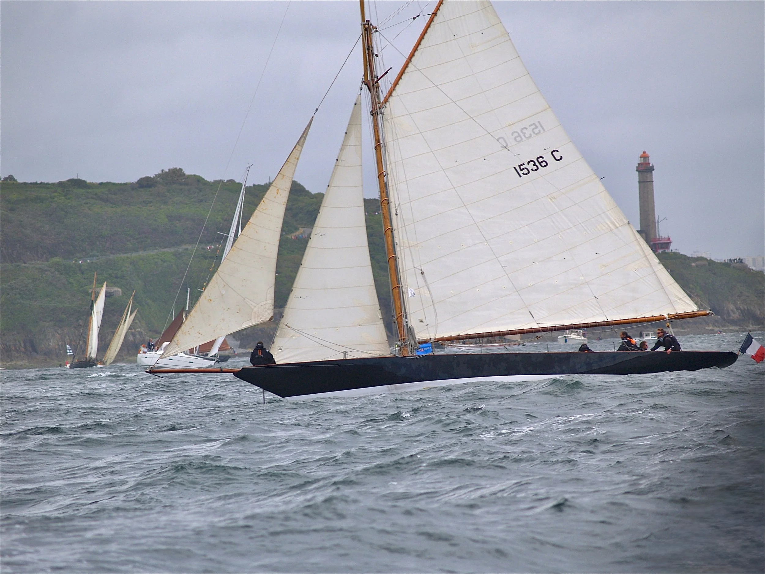 Voilier avec des voiles en toile blanche naviguant sur la mer avec une côte verdoyante en arrière-plan et un phare visible sur la droite