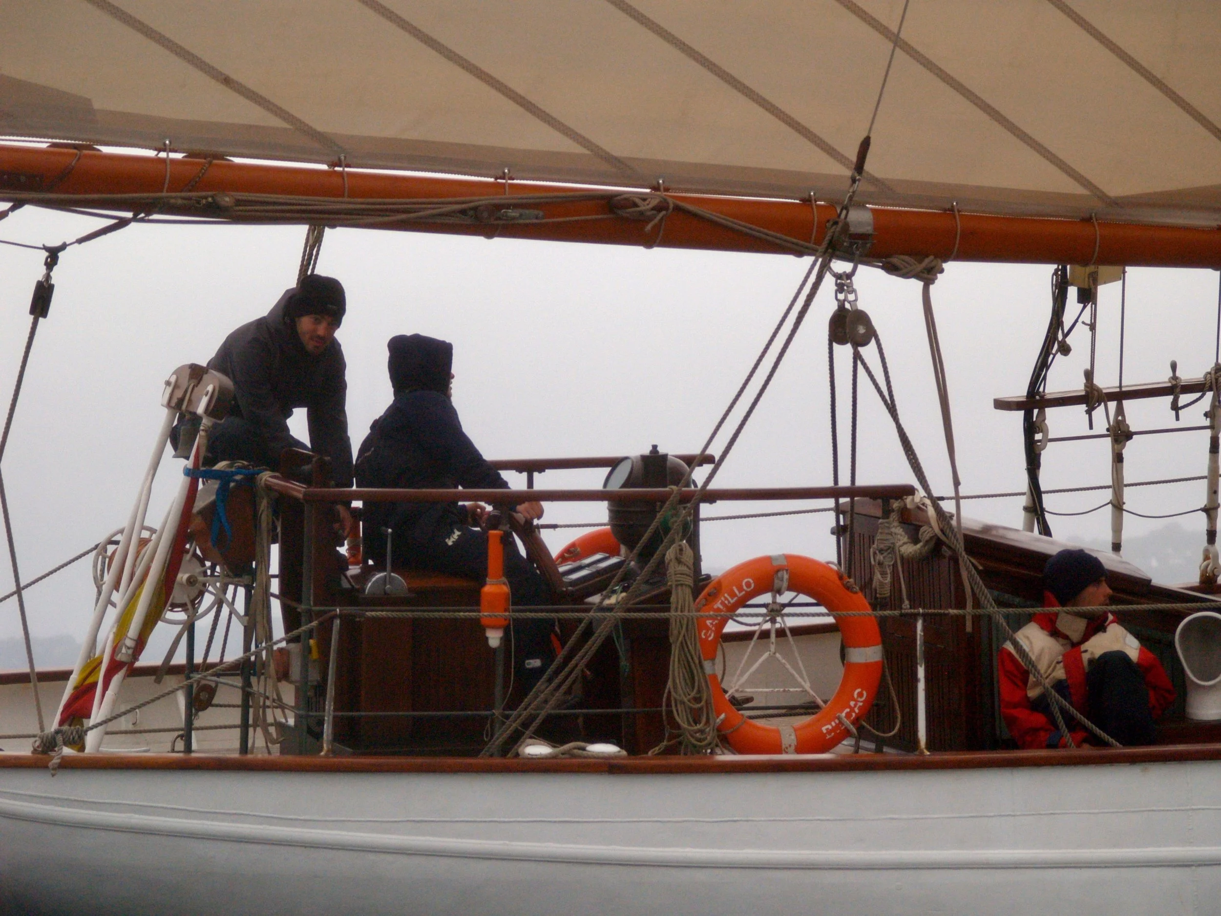 Vue de l'intérieur d'un bateau avec deux hommes discutant, divers cordages et équipements de navigation, et une vie sauvetage orange visible.