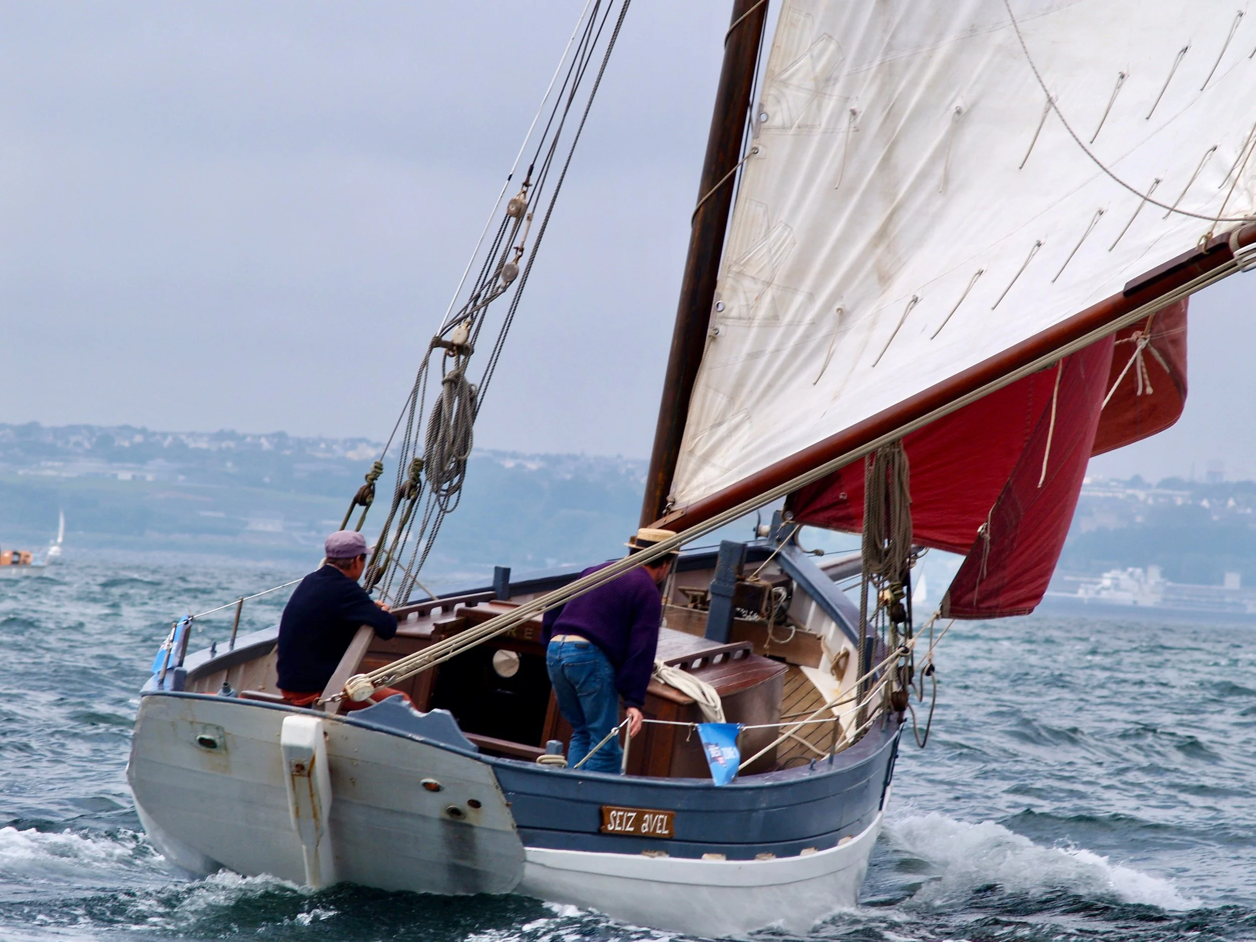 Un voilier naviguant sur l'eau avec deux personnes à bord, un des voiliers porte une voile blanche et une coque bleue. Il y a un paysage côtier en arrière-plan.