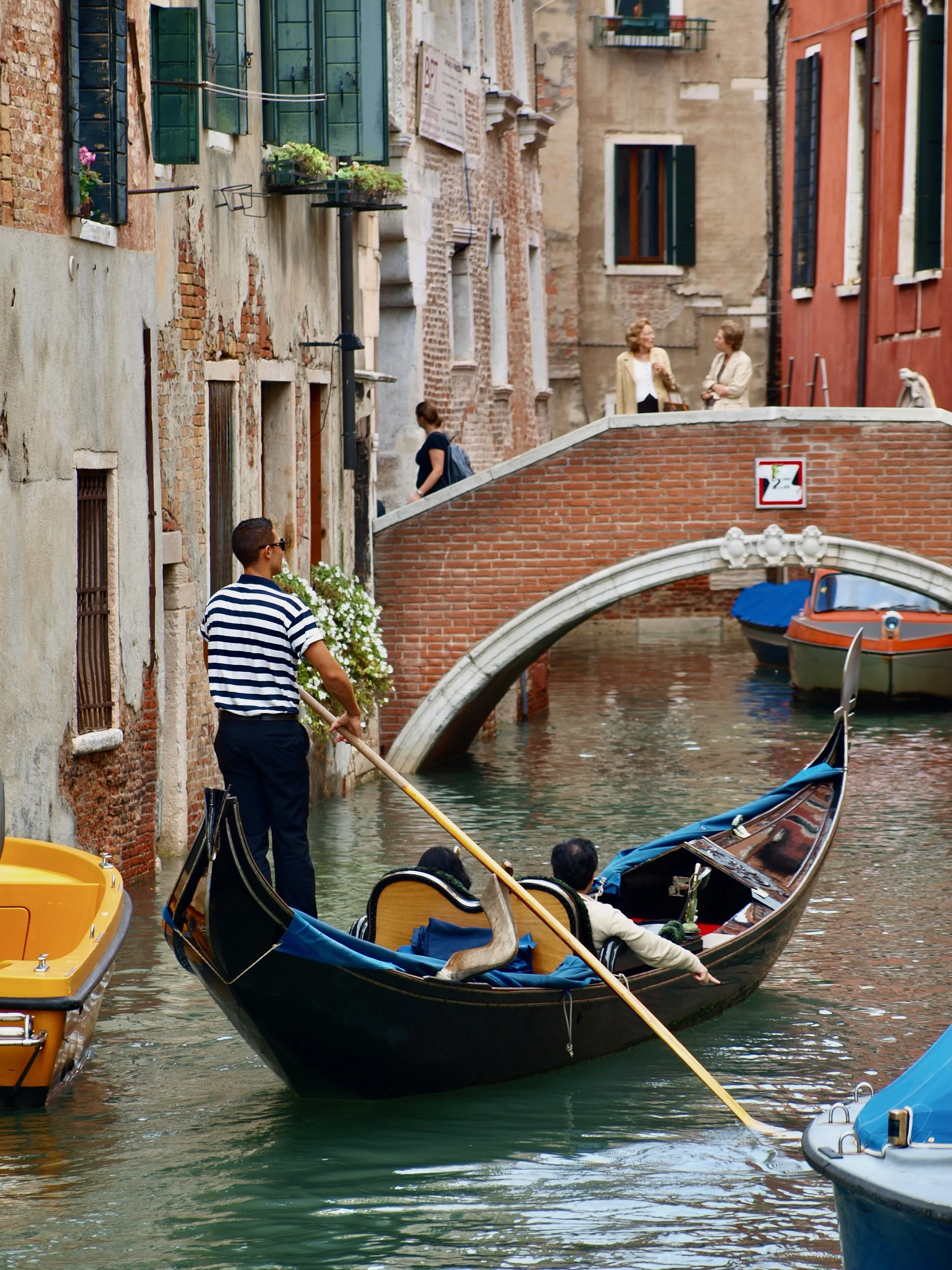 Une scène de canal à Venise avec des gondoles, une femme en uniforme de gondolier, et des bâtiments anciens avec des fenêtres à volets verts et des plantes en fleurs