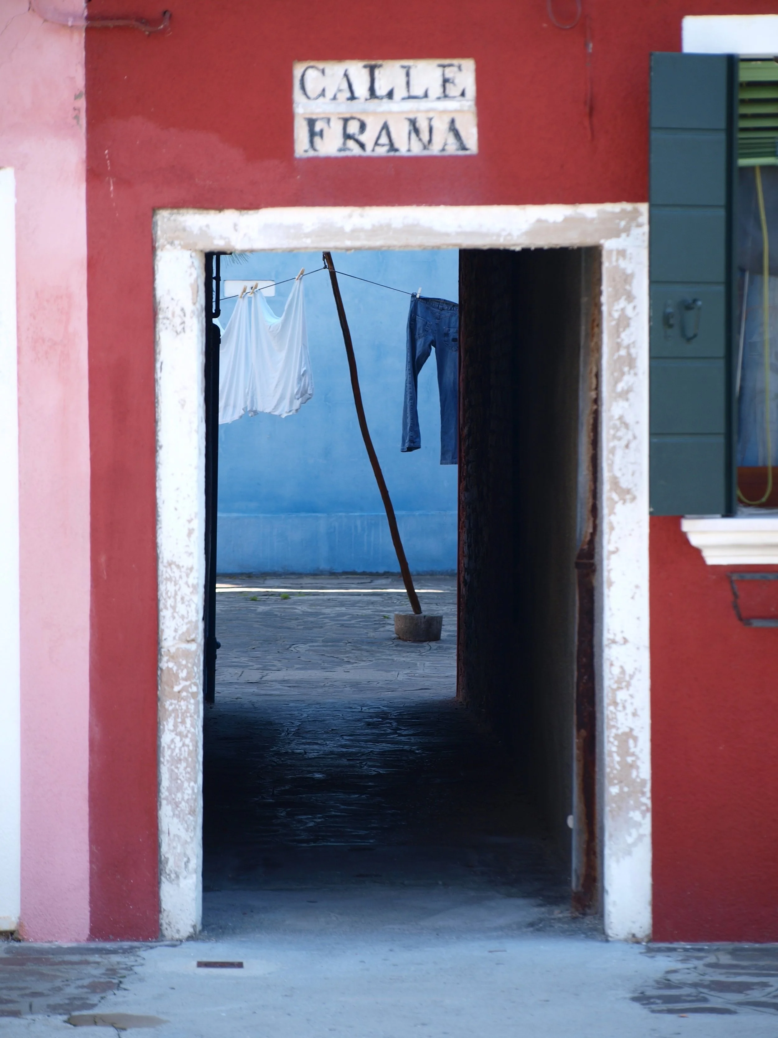 Vue à travers une porte vers une cour avec linge suspendu pour sécher, entourée de murs colorés, avec un panneau indiquant 'Calle Frana'