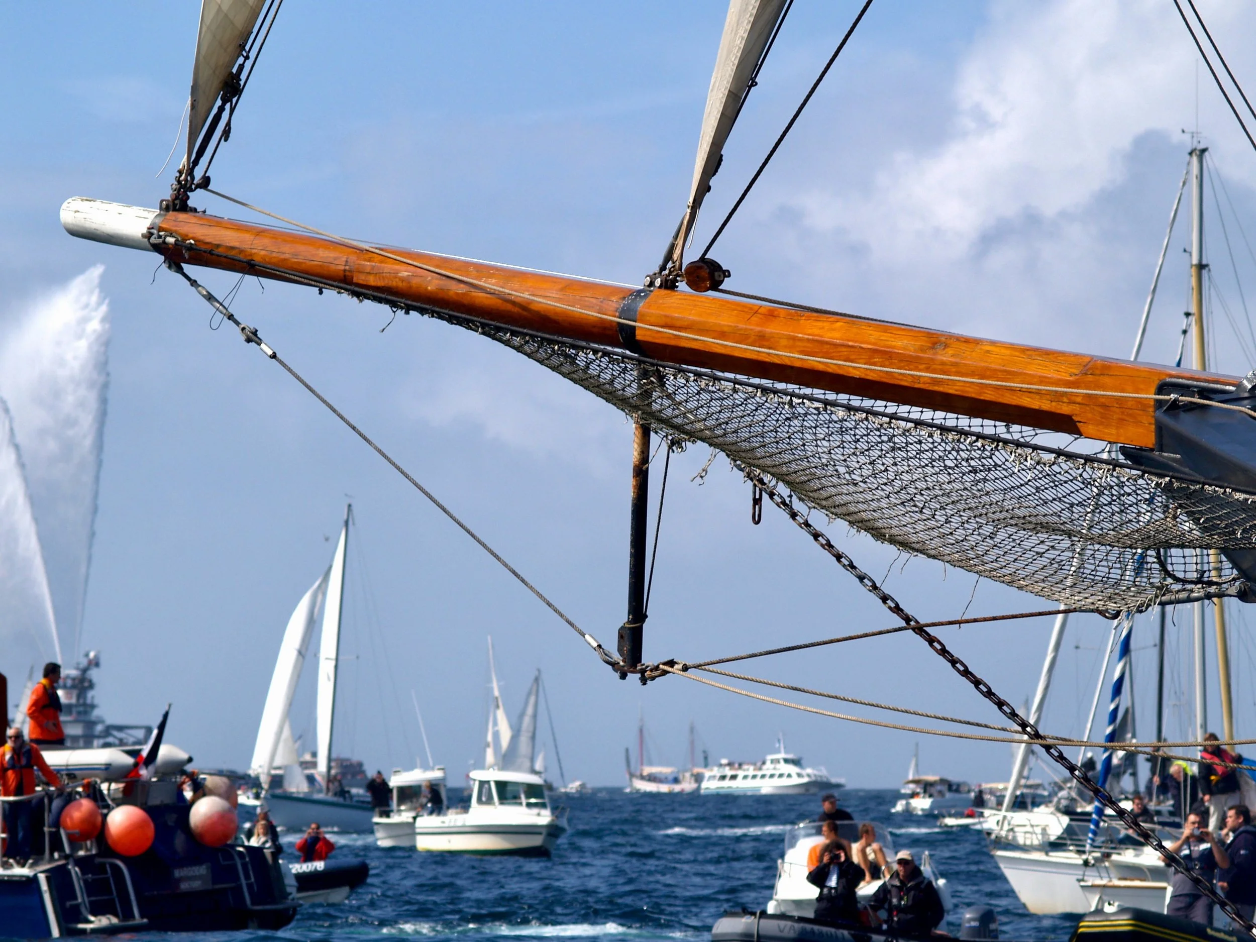 Voile en bois avec des cordages, dans un port rempli de bateaux à voile modernes et de plaisance, avec des personnes sur le quai, sous un ciel partiellement nuageux.