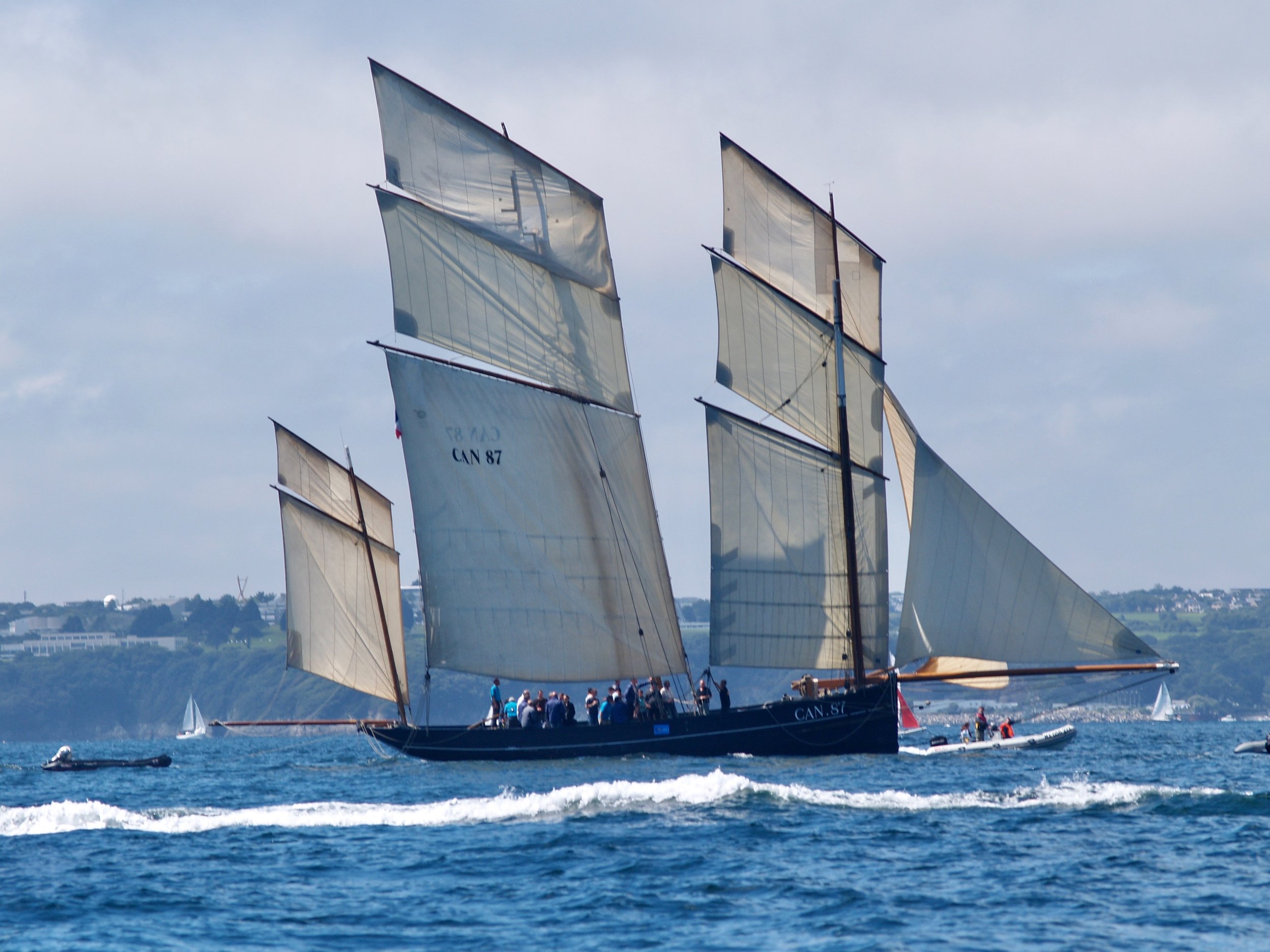 Bâteau à voile traditionnel avec plusieurs voiles déployées naviguant sur l'eau, avec des spectateurs à bord et en radeau, sous un ciel partiellement nuageux.