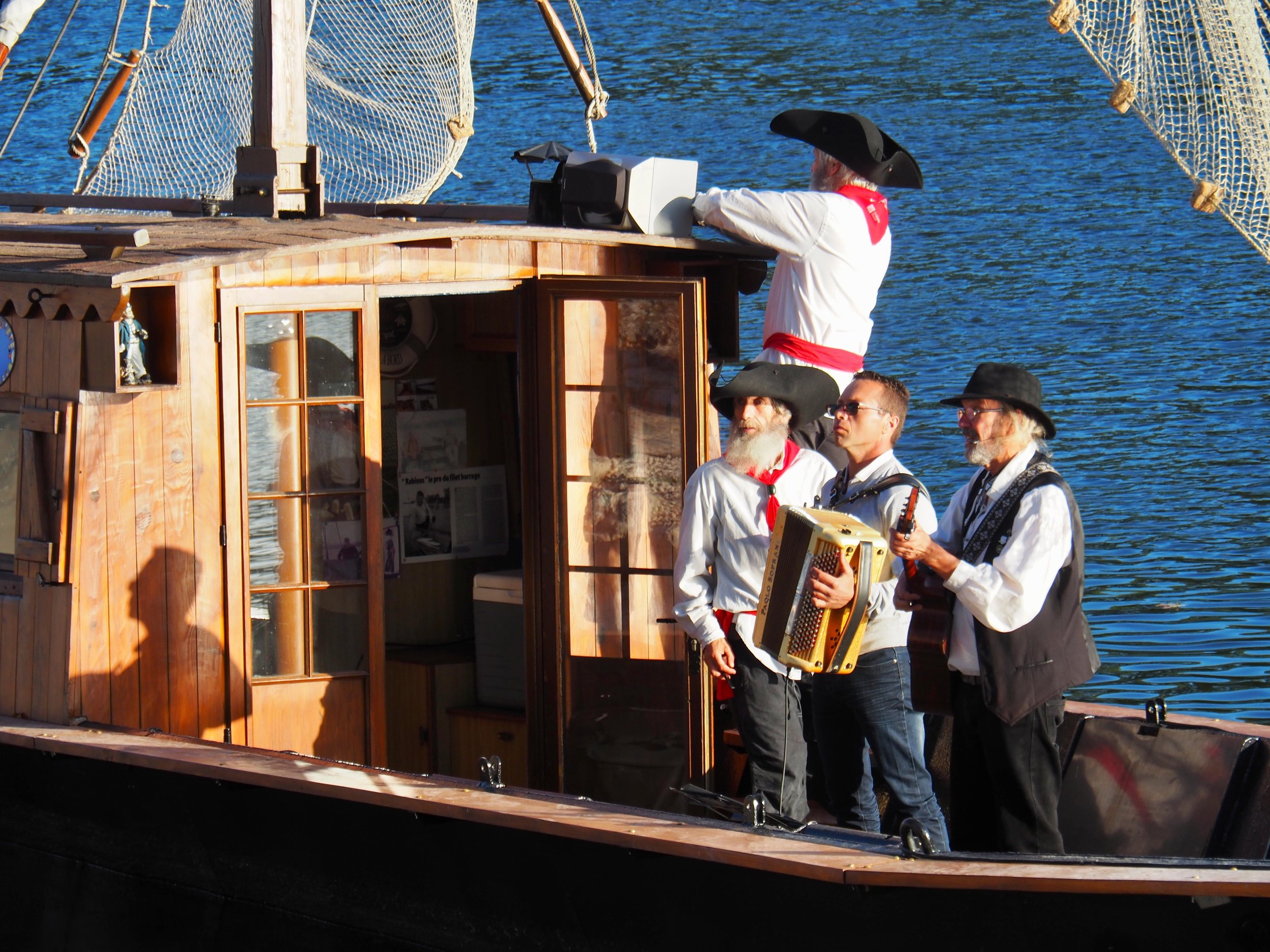 Groupe de personnes habillées en costume traditionnel, avec certains portant des chapeaux, sur un bateau en bois en mer. La scène est en plein jour avec de l'eau bleue en arrière-plan.