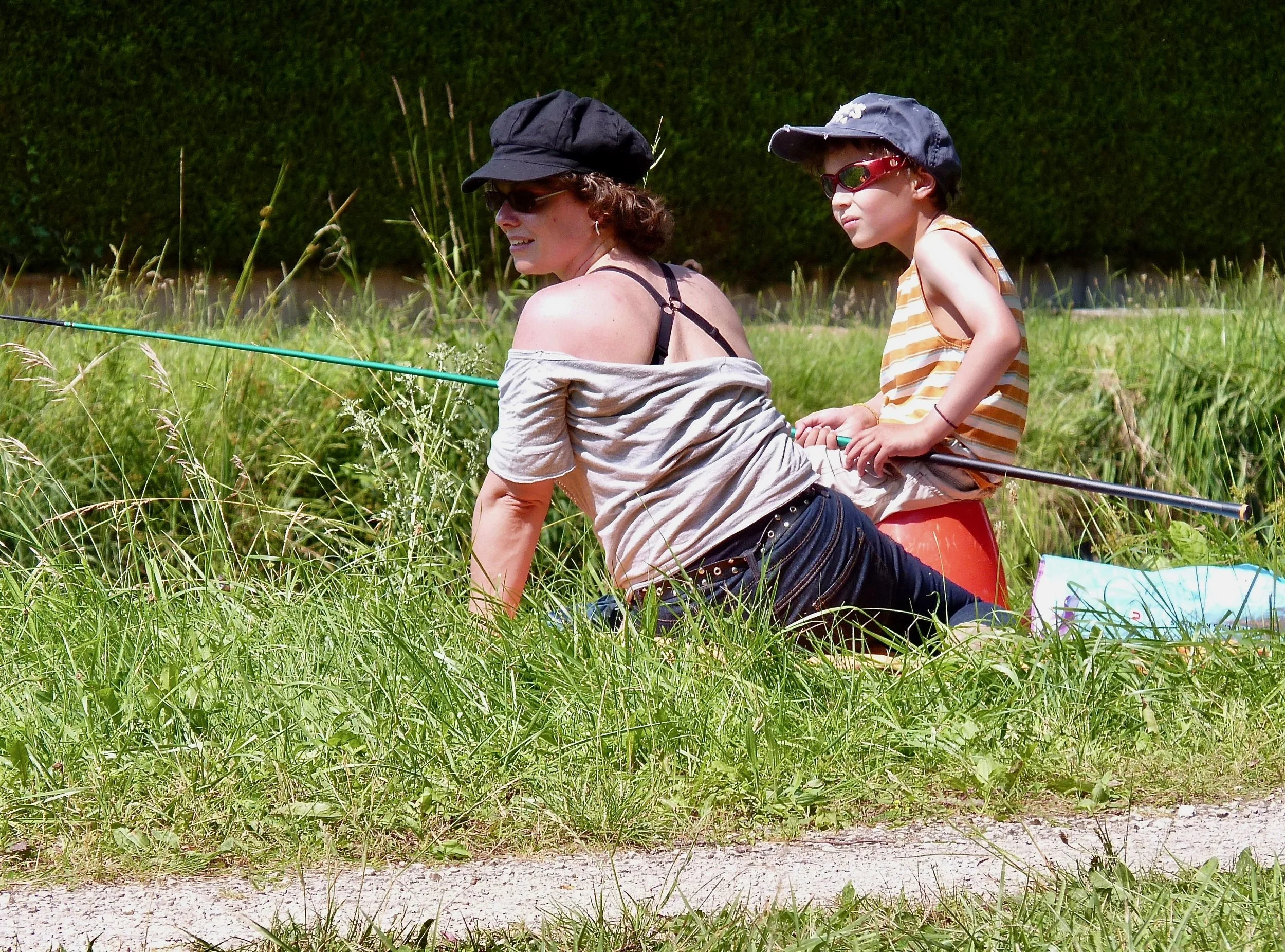 Une femme et un enfant faisant du camping ou de la pêche dans un espace extérieur vert avec de hautes herbes, le soleil en journée, tous deux portant des chapeaux et des lunettes de soleil, l'enfant assis dans un siège ou un support, tous deux concen