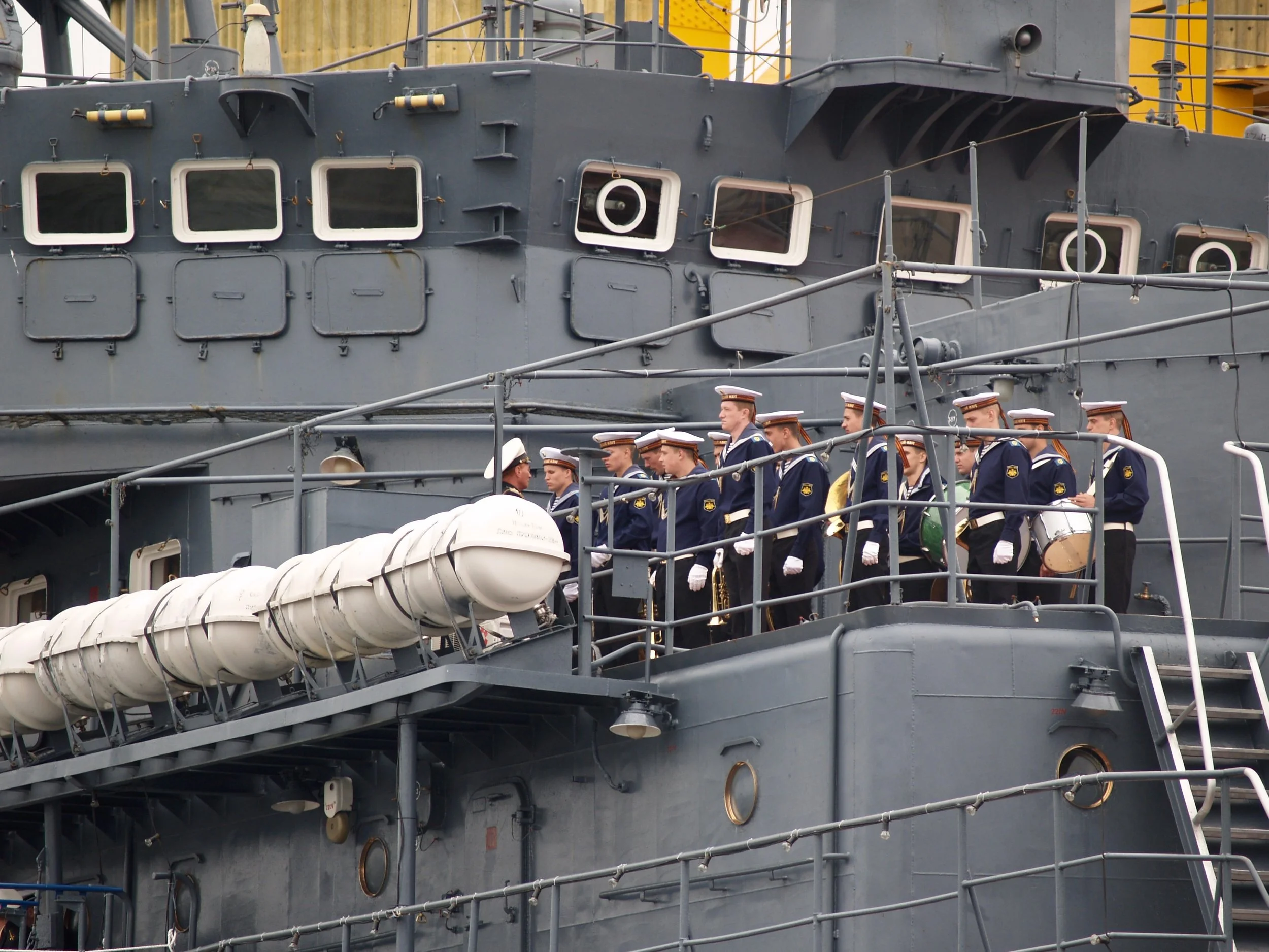 Une scène de cérémonie militaire avec des soldats en uniformes blancs marchant sur le pont supérieur d'un grand navire de guerre avec une superstructure en acier gris, plusieurs fenêtres, équipements et cordages.