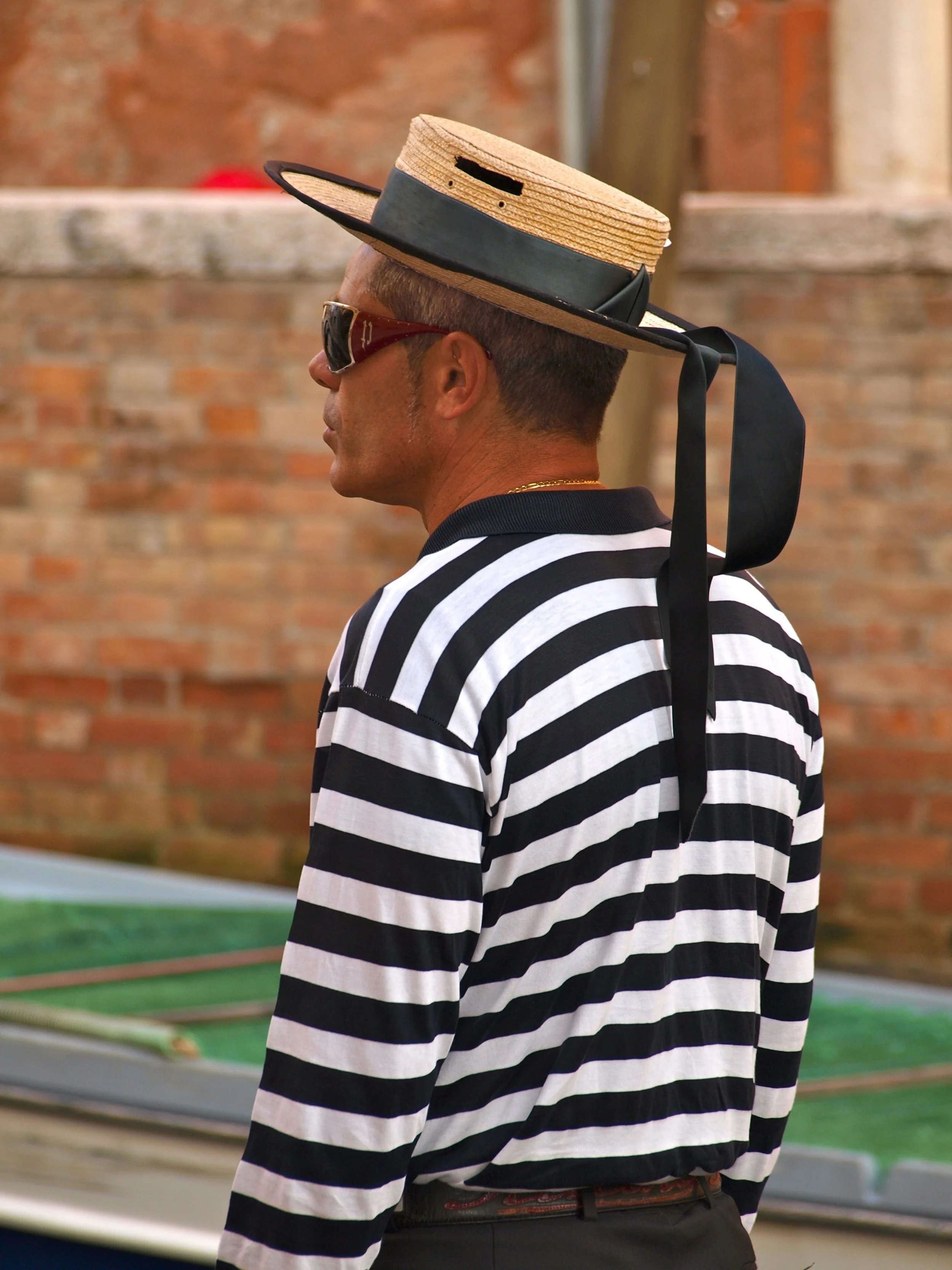 Homme portant un chapeau en paille avec un ruban noir, lunettes de soleil, et un maillot à rayures noires et blanches, à l'extérieur devant un mur en briques.