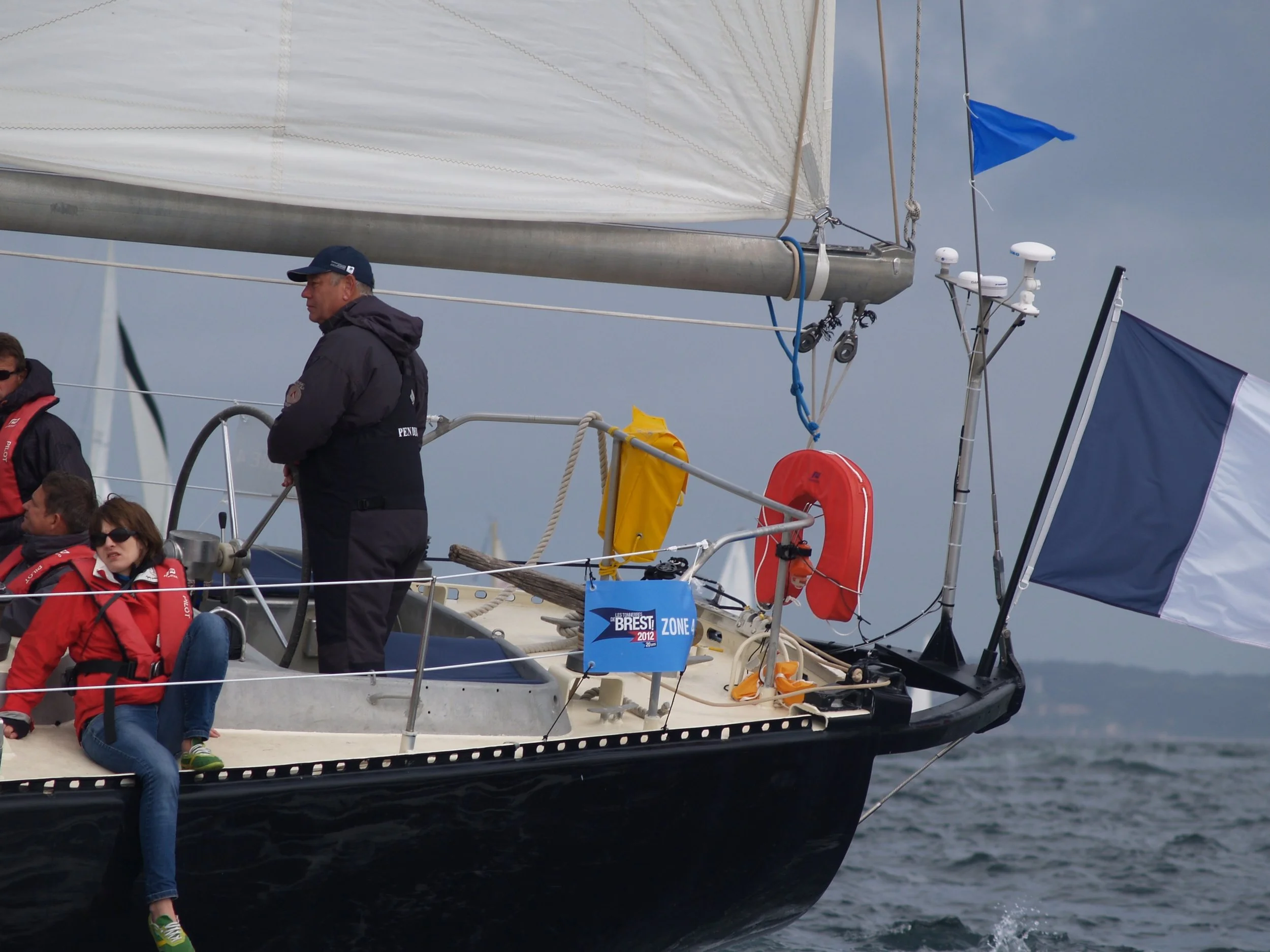 Groupe de personnes à bord d'un voilier, avec certains assis et d'autres debout, portant des vêtements de voile dans une journée nuageuse.