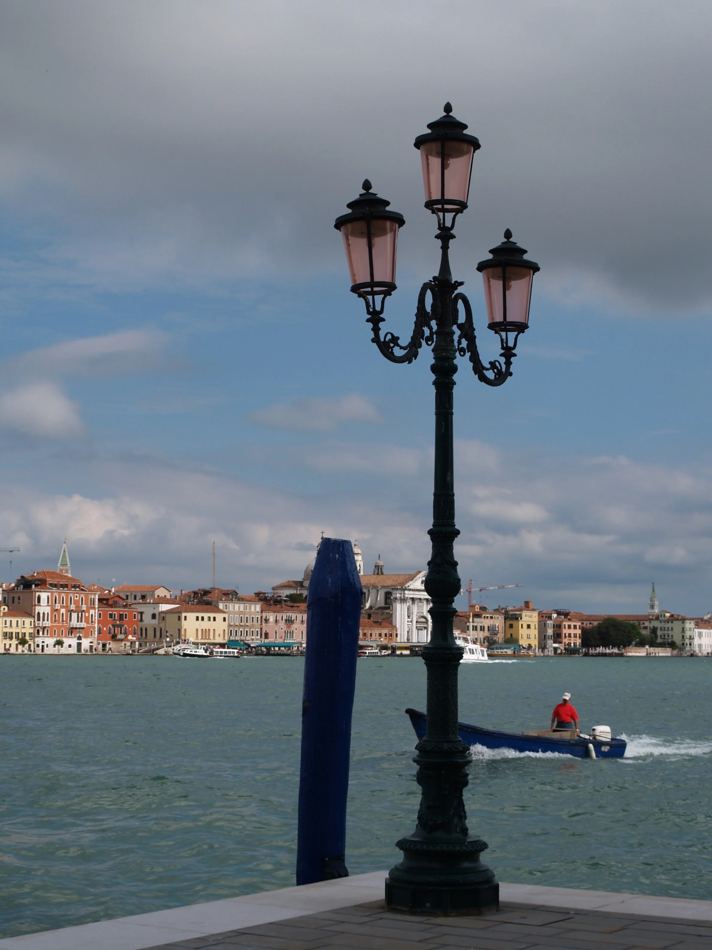 Une lampe de style ancien au premier plan avec un fond de ville côtière et de l'eau, un bateau naviguant dans la baie, et un ciel partiellement nuageux.