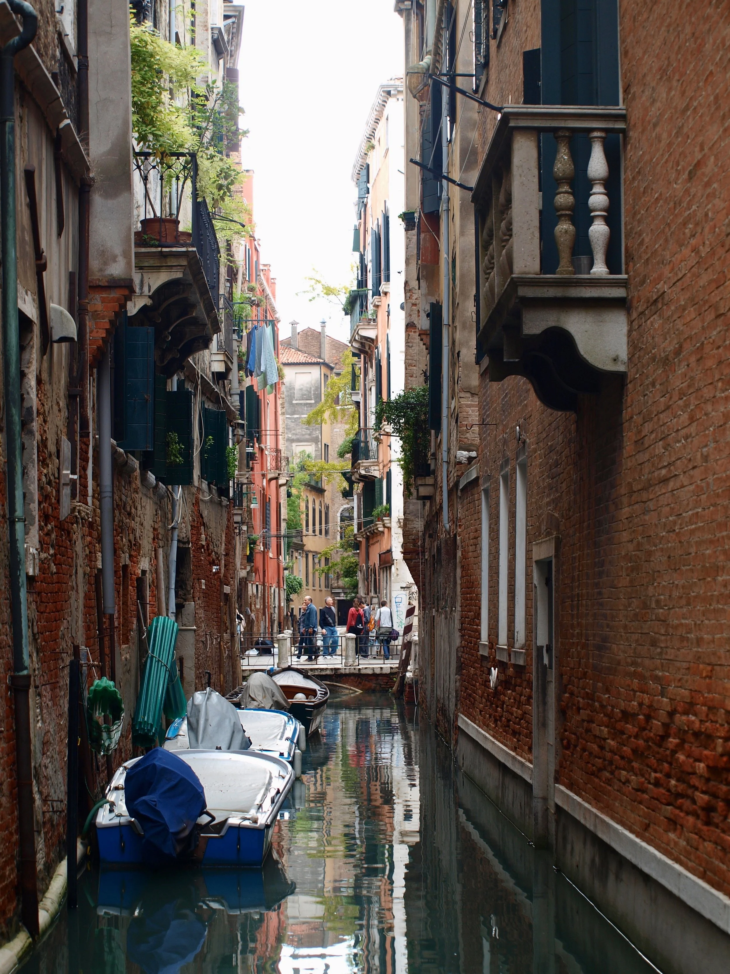 Ruelle étroite bordée de bâtiments en briques avec des balcons fleuris et des volets bleus, vue d'une petite embarcation dans un canal, à Venise.