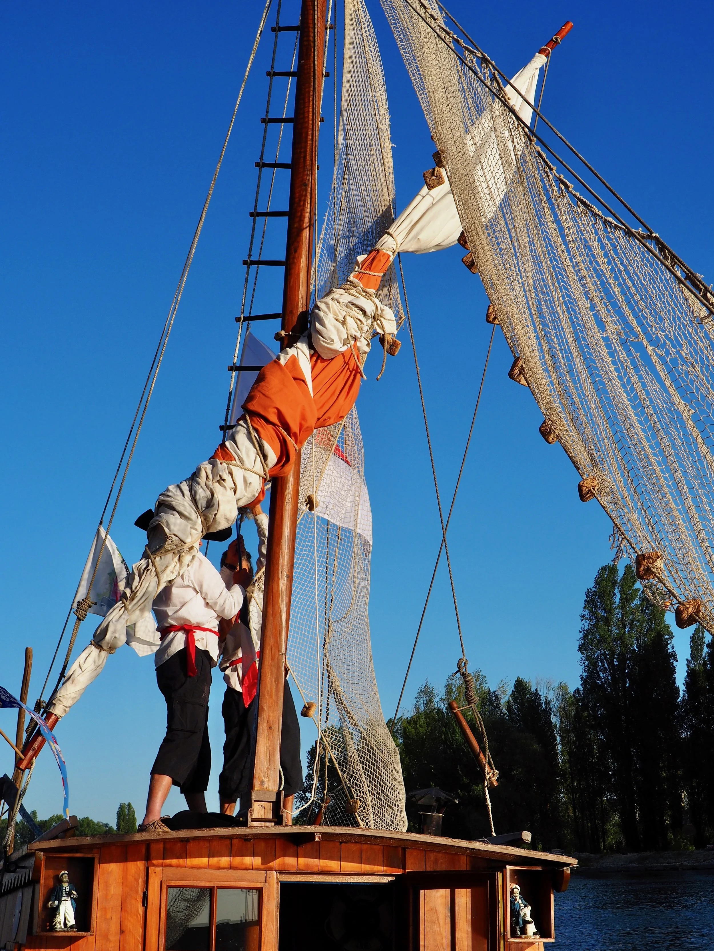 Un voilier en bois avec des voiles en coton, des filets et une échelle en bois, sous un ciel bleu clair, avec des personnes en tenue de marin faisant partie de l'équipage.