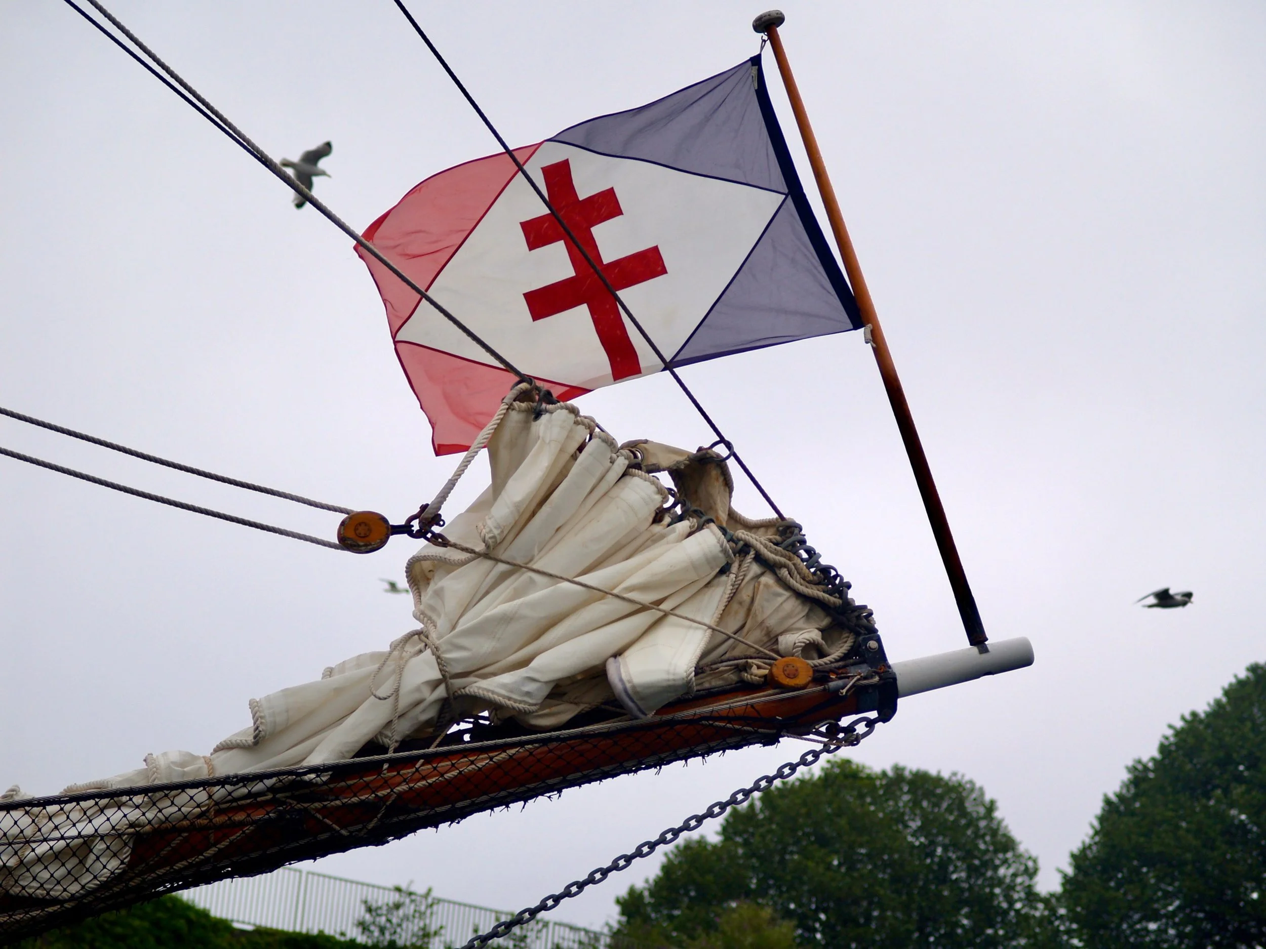 Drapeau de la Croix-Rouge flottant au sommet d'une voile de bateau, avec des nuages gris et des oiseaux en arrière-plan.