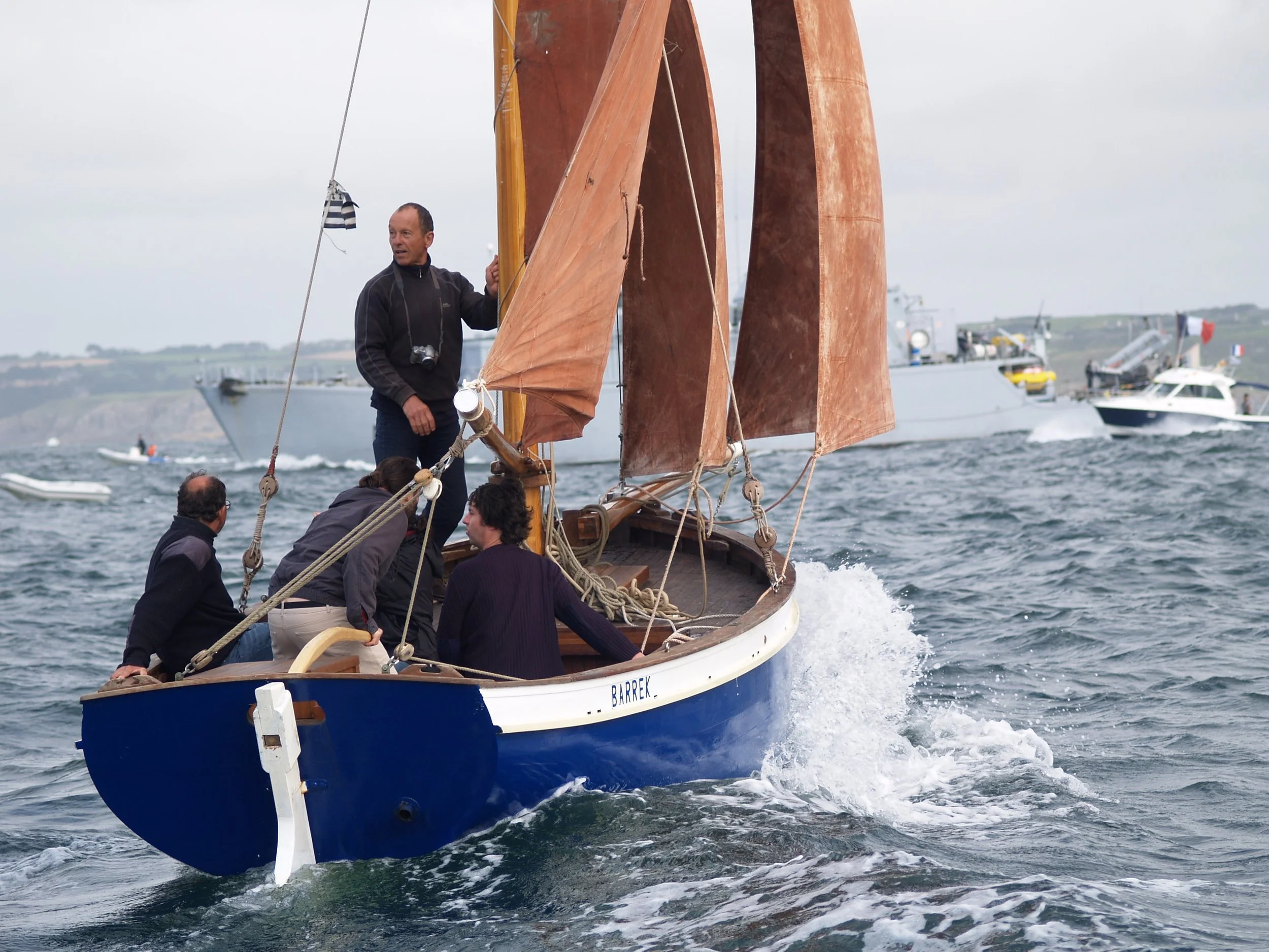 Un groupe de personnes naviguent sur un bateau en bois avec des voiles en toile de jute tandis que des bateaux modernes naviguent en arrière-plan en mer par un jour nuageux.