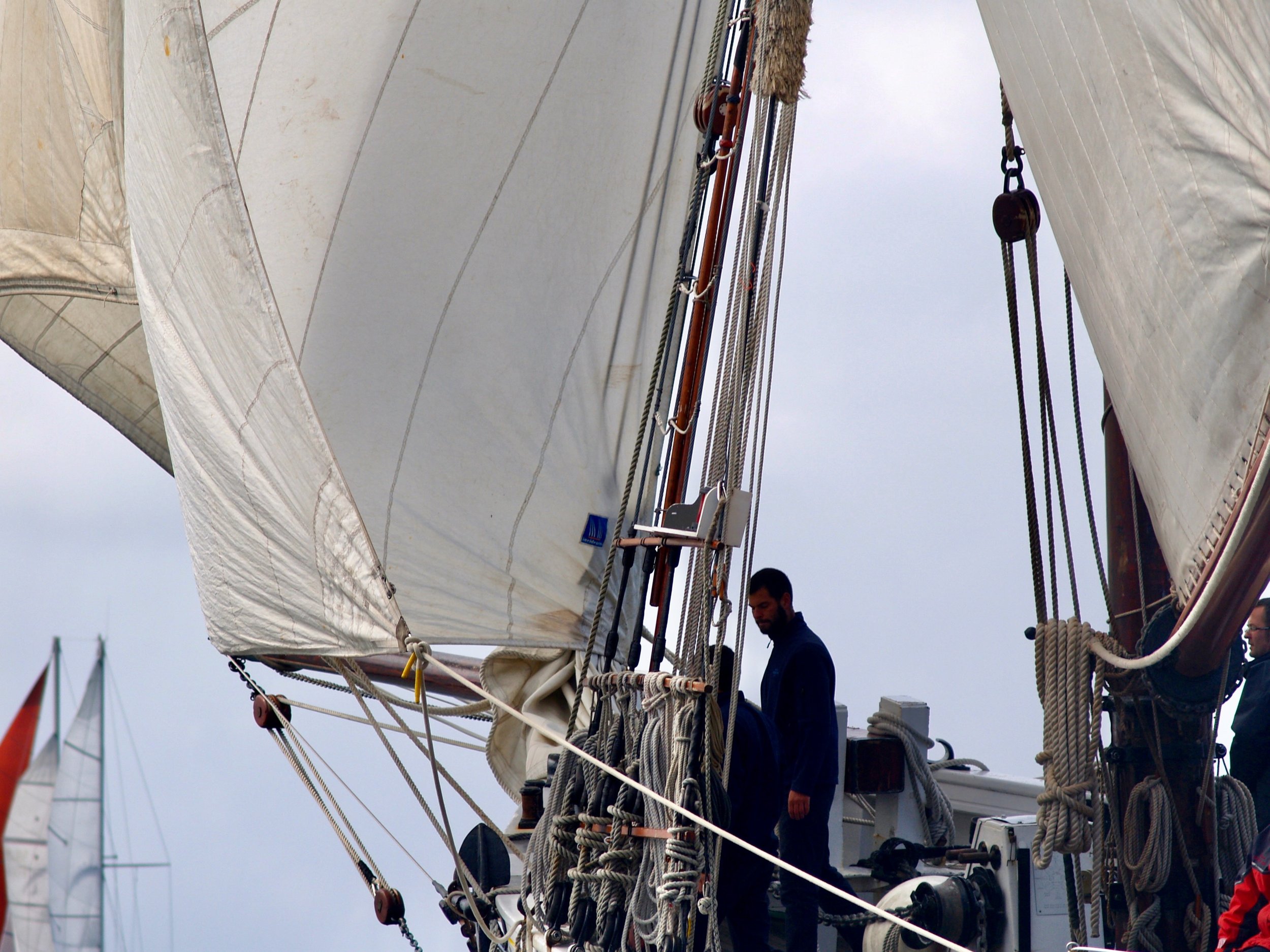 Gens en tenue de marin à bord d'un voilier, entourés de cordages et manœuvres, sous un ciel nuageux.